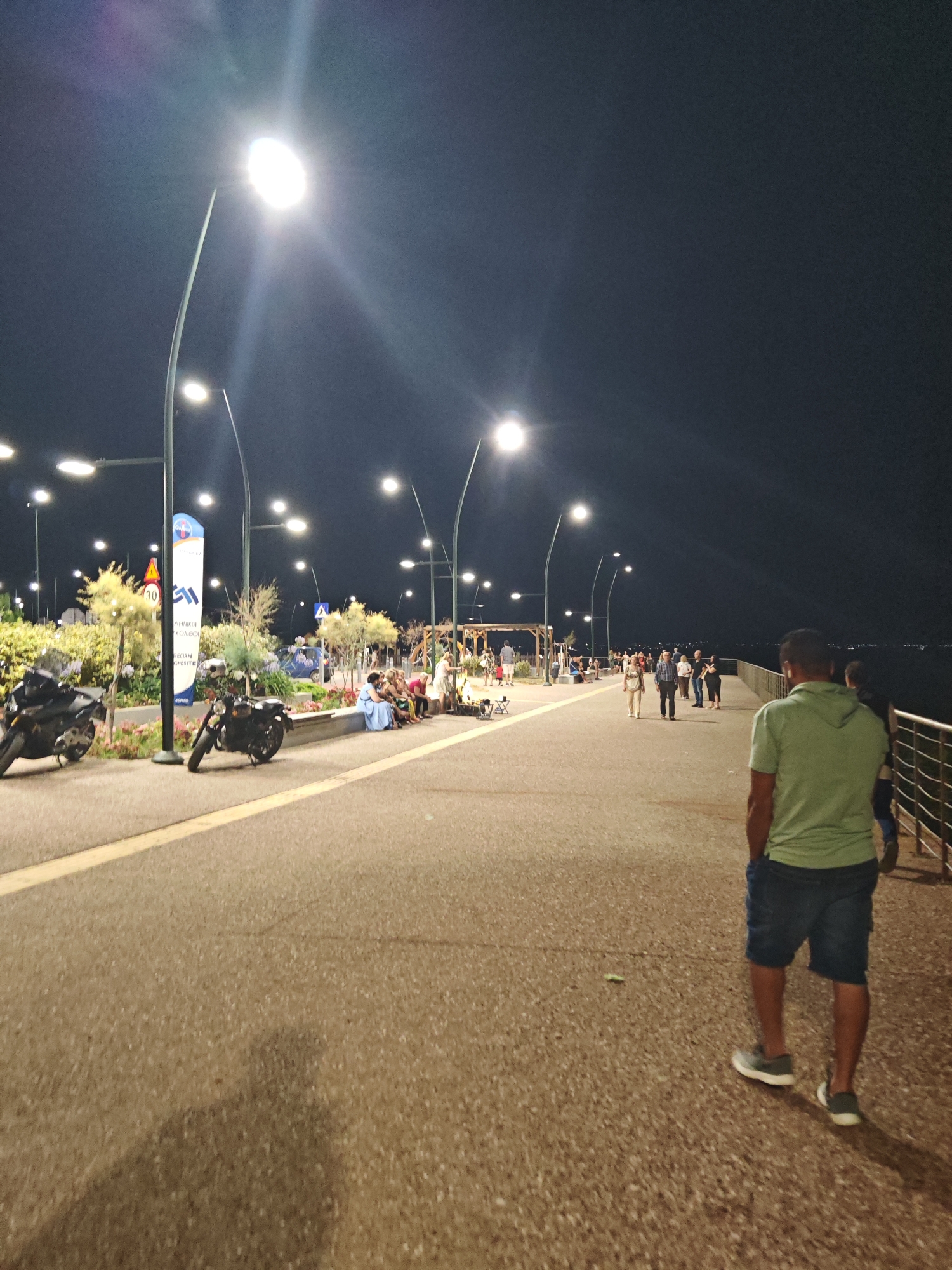 The image shows a nighttime scene on a waterfront promenade. A man in a green t-shirt and blue shorts walks away from the camera on the right side of the frame. The promenade is lined with streetlights and a metal railing separates it from the water on the right. On the left side of the promenade, there are parked motorcycles, a seating area with several people sitting, and landscaping. In the background, more people are walking along the promenade. The sky is dark and clear, with a few lights visible in the distance over the water.