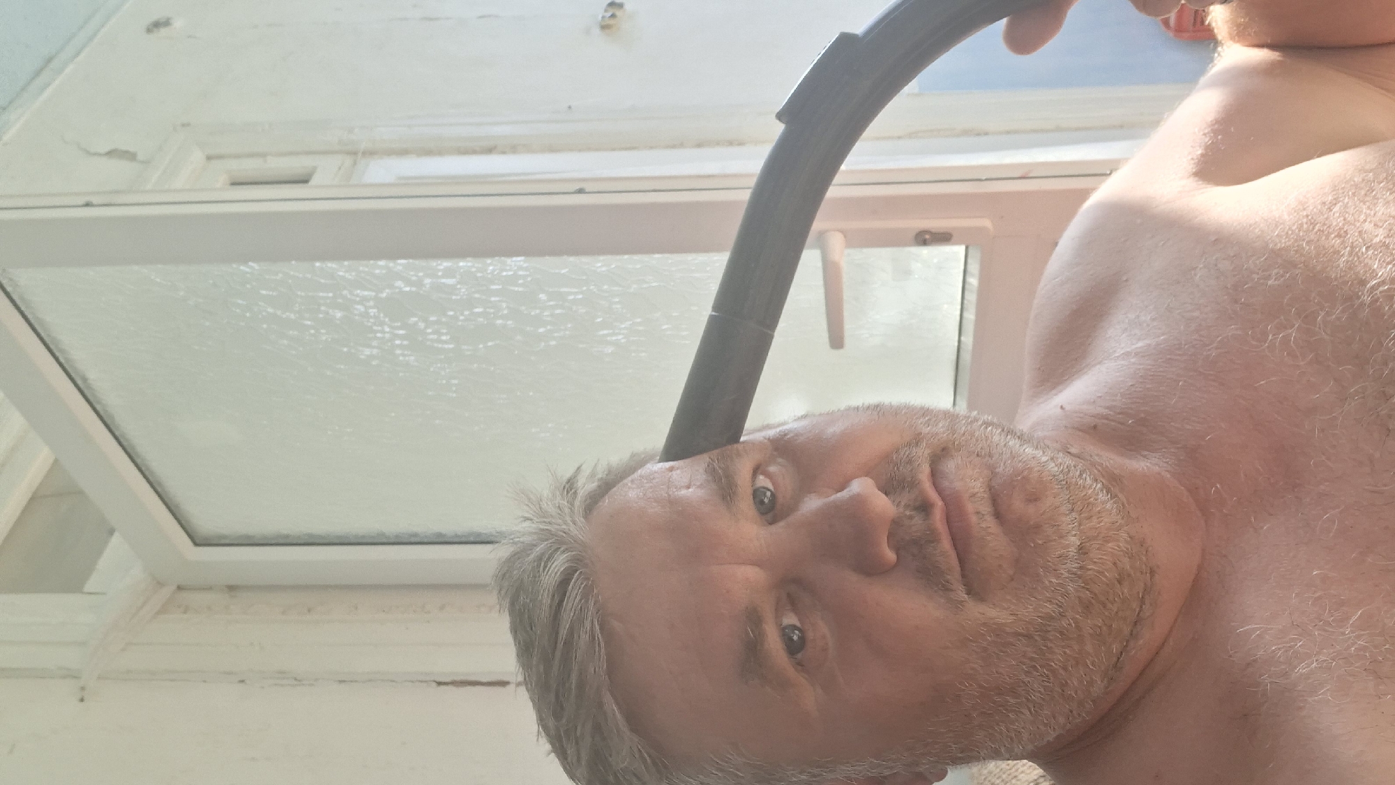 A close-up, selfie-style shot features a shirtless middle-aged man with grey hair and a beard. He is positioned in front of a partially frosted glass door and a white wooden wall, suggesting an indoor or semi-outdoor setting. A black, curved hose or pipe is held against the right side of his head, obscuring part of his face and extending out of frame. The man's expression is neutral, and his gaze is directed towards the camera. The lighting is bright, casting shadows and highlighting textures on his skin and the surrounding environment. The scene appears to be in Voinova, Moldova, based on the provided context, but there are no specific visual cues within the image itself to confirm this location.