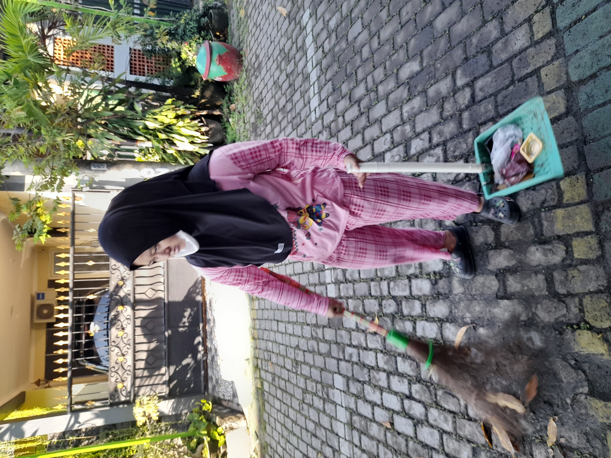 A person is sweeping a cobblestone driveway or walkway with a broom in Gelam, Indonesia. The person wears a black hijab, a pink long-sleeved shirt with a butterfly print, and pink plaid pants. They also wear black clog-style shoes. The person is using a broom with a green handle and a brown bristle head to sweep debris on the cobblestones, and is holding a dustpan with a light green frame and garbage inside. The driveway or walkway leads toward a house with a black iron gate. Trees and potted plants are located on the side of the house.
