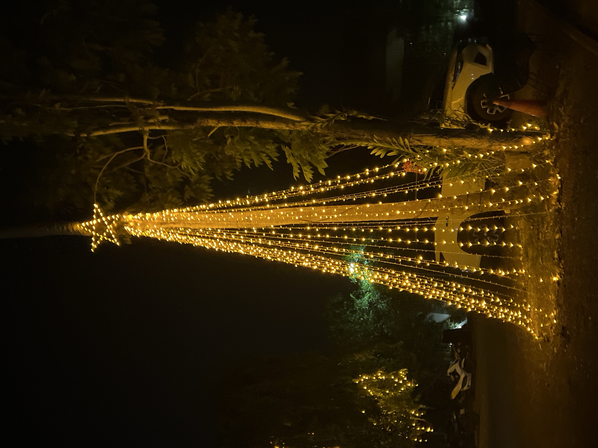This image depicts a nighttime scene in Ban Bang Rak, Thailand, decorated with a multitude of warm yellow string lights. The lights are strung in a cascading manner from a tree, forming a triangular shape reminiscent of a Christmas tree. A star-shaped light fixture is visible at the top of this illuminated structure. In the background, a white SUV is parked, partially obscured by branches and greenery. The ground appears to be a dirt or gravel surface. The overall ambiance is festive and enchanting, suggesting a special occasion or holiday celebration. The time of day is clearly night, and the lighting creates a magical atmosphere.
