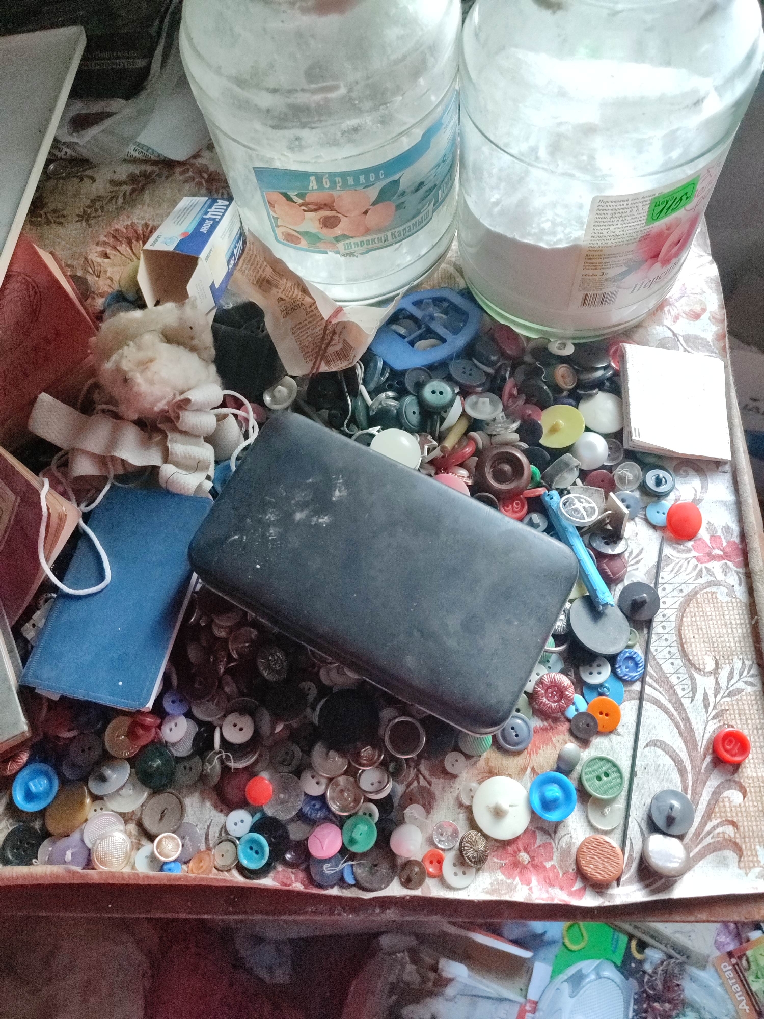 The image depicts a cluttered tabletop scene, possibly in an apartment in Gremyachinsk, Russia. Dominating the top of the frame are two large, clear glass jars, both containing a white, powdery substance, likely flour or sugar, with labels suggesting contents like "Apricot" and "Percussion." Below the jars, a multitude of colorful buttons are scattered across a patterned tablecloth. These buttons vary in size, shape, and material, with some appearing to be made of plastic and others of shell or metal.
In the center of the scene, a dark, rectangular object, possibly a jewelry box or a similar container, rests atop a pile of these buttons. To the left of this object, a small, blue-covered notebook and a red-covered book are visible, along with some rolled-up white fabric and a tuft of light-colored material. The overall impression is one of disarray and a collection of crafting or household items. The lighting is somewhat dim, suggesting an indoor environment. There are no people or explicit actions taking place in the image.