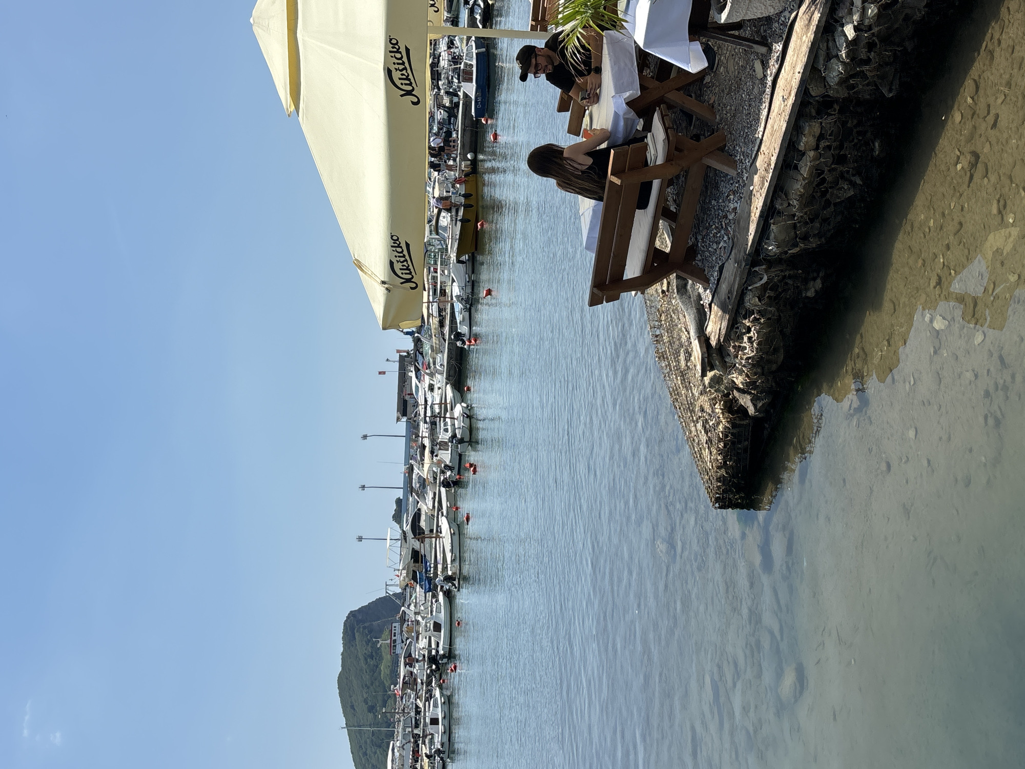 The image depicts a sunny day at a harbor in Budva, Montenegro. Two people, a man and a woman, are seated at a table under a large, light-colored umbrella, appearing to be enjoying a meal or drinks. The man, wearing a black cap and glasses, leans forward towards the table. The woman, with long dark hair, is also looking down at the table. The setting is a waterfront restaurant with wooden tables and benches.
In the background, a marina is filled with numerous white boats of varying sizes, docked along the water. The calm, blue water reflects the clear sky and the boats. A verdant, mountainous landscape rises in the distance. The time of day appears to be late morning or early afternoon, given the bright sunlight and clear skies. The overall mood is relaxed and leisurely, suggesting a pleasant vacation atmosphere.
Visible text on the umbrella reads "Nikšićko" in black lettering. The ground around the restaurant appears to be a mix of gravel and stone. The water's edge is defined by a stone embankment.