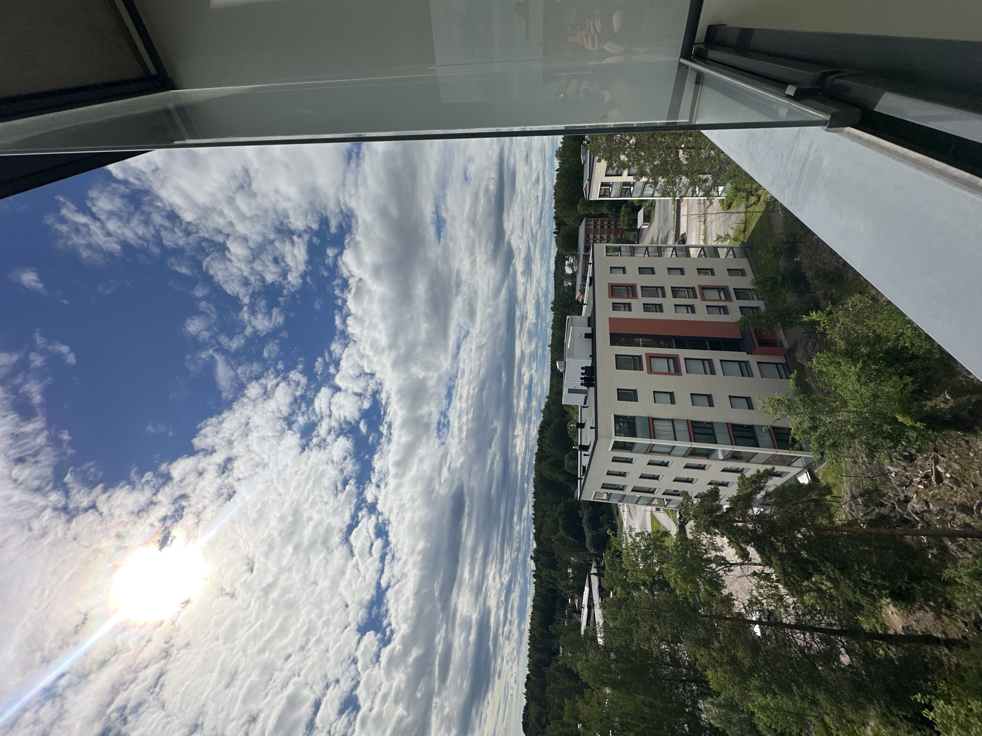 The image is a vertical, wide-angle view of an urban landscape with a focus on architecture and nature. In the foreground, a modern, multi-story apartment building with white and light orange accents and numerous windows dominates the right side of the frame. Trees and greenery surround the building, with some rocky terrain visible. Beyond the apartment building, more trees and a road can be seen, suggesting a residential area.
The left side of the image is dedicated to the sky. A bright sun, with a visible lens flare, shines through a dramatic sky filled with a mix of bright white, fluffy clouds and darker, streaky clouds. The blue sky peeks through in areas, creating a dynamic contrast. The angle suggests the photo was taken from a high vantage point, possibly a balcony or window of another building.
The overall impression is a sunny day with a mix of weather conditions, evidenced by the varied cloud formations. The scene conveys a sense of a modern living environment integrated with natural elements. There is no visible text in the image. The time of day appears to be daytime, given the bright sunlight.