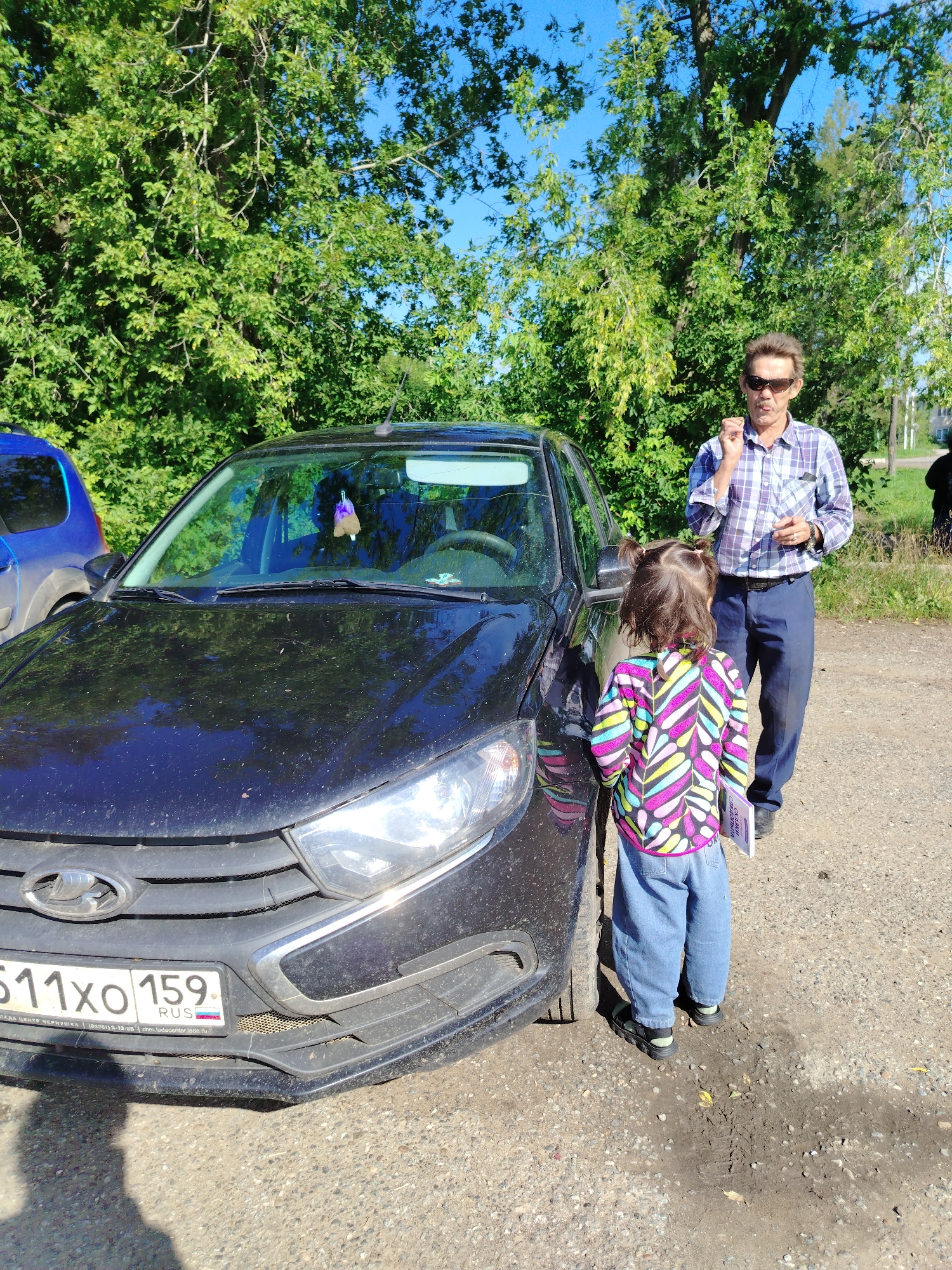 A medium-shot photograph captures a scene outdoors, likely on a sunny day, with a black car parked on a gravel surface. Behind the car, a dense line of green trees forms a natural backdrop under a clear blue sky.
In the foreground, a young child, seen from the back, stands beside the car. The child has dark hair styled into two pigtails and is wearing a brightly colored, striped jacket with a zig-zag pattern in shades of pink, yellow, blue, and green, along with loose-fitting blue jeans and black sandals. The child is holding a thin book or pamphlet.
To the right of the child, a man stands facing the child and the car. He is wearing sunglasses, a blue and white plaid collared shirt, and dark blue pants. His right hand is raised to his mouth, and he appears to be holding a small object between his fingers. He is also holding a white object in his left hand.
The black car is partially visible, showing its front left side. The license plate is clearly legible: "511XO 159" with "RUS" below it. The car's headlights and grille are also visible. To the left of the black car, the rear of a blue vehicle is partially visible.
The ground is covered in small stones and dirt, with some scattered leaves and muddy patches indicating recent moisture. The overall setting suggests a rural or semi-rural environment, possibly a parking area near a natural space. Based on the Russian license plate, the location is in Russia, and the license plate format is consistent with registration in the Kuyeda region of Russia.