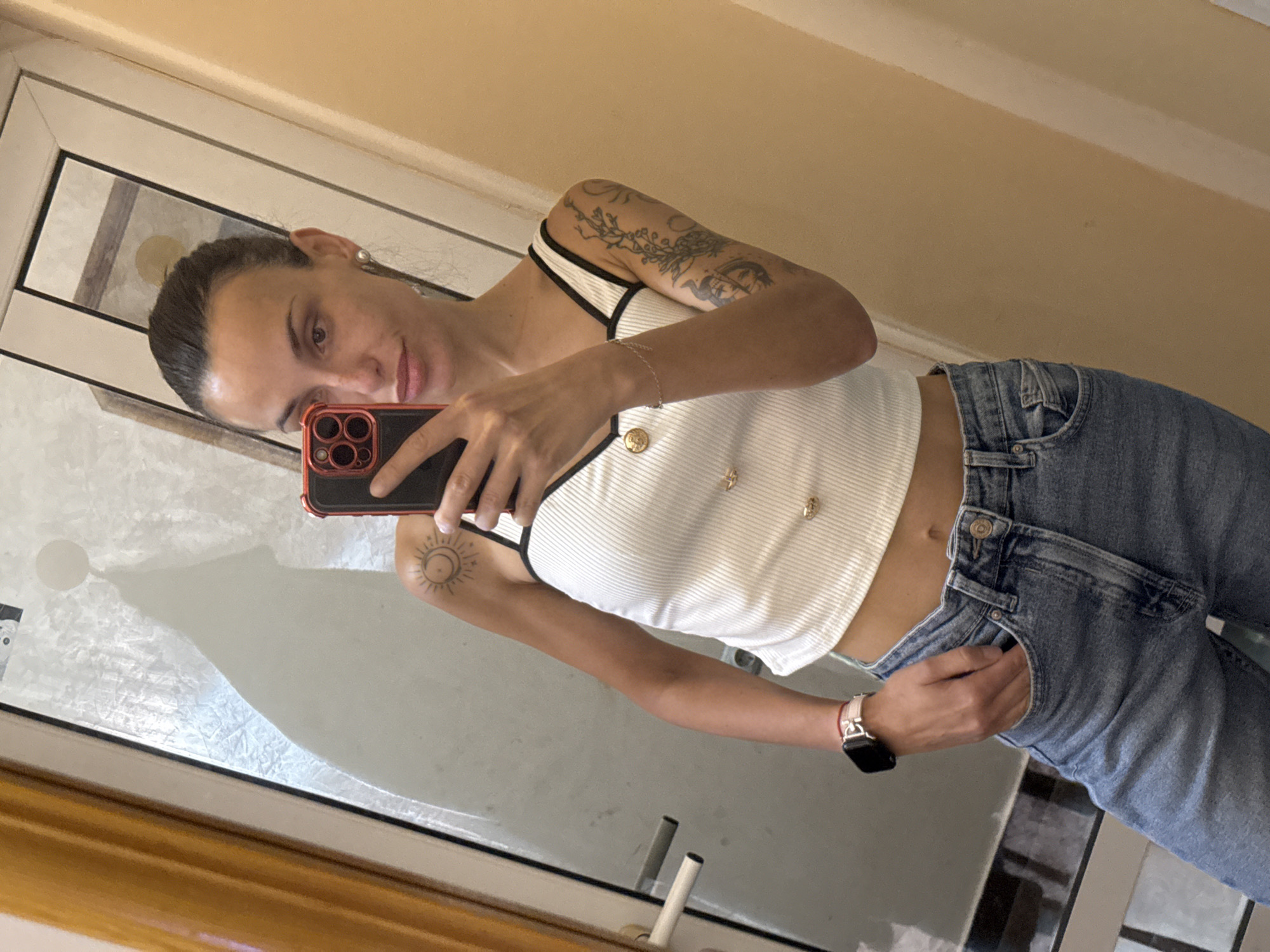 A woman stands in front of a mirror, holding a smartphone in her right hand to take a selfie. She is wearing a white, ribbed tank top with black straps and blue jeans. Her left hand is partially in her jeans pocket. The woman has a tattoo on her left forearm and another on her right upper arm. She is wearing an Apple Watch on her left wrist. The background includes a door with decorative molding. The location is likely indoors, but there are no details to link this image to Pleven, Bulgaria.