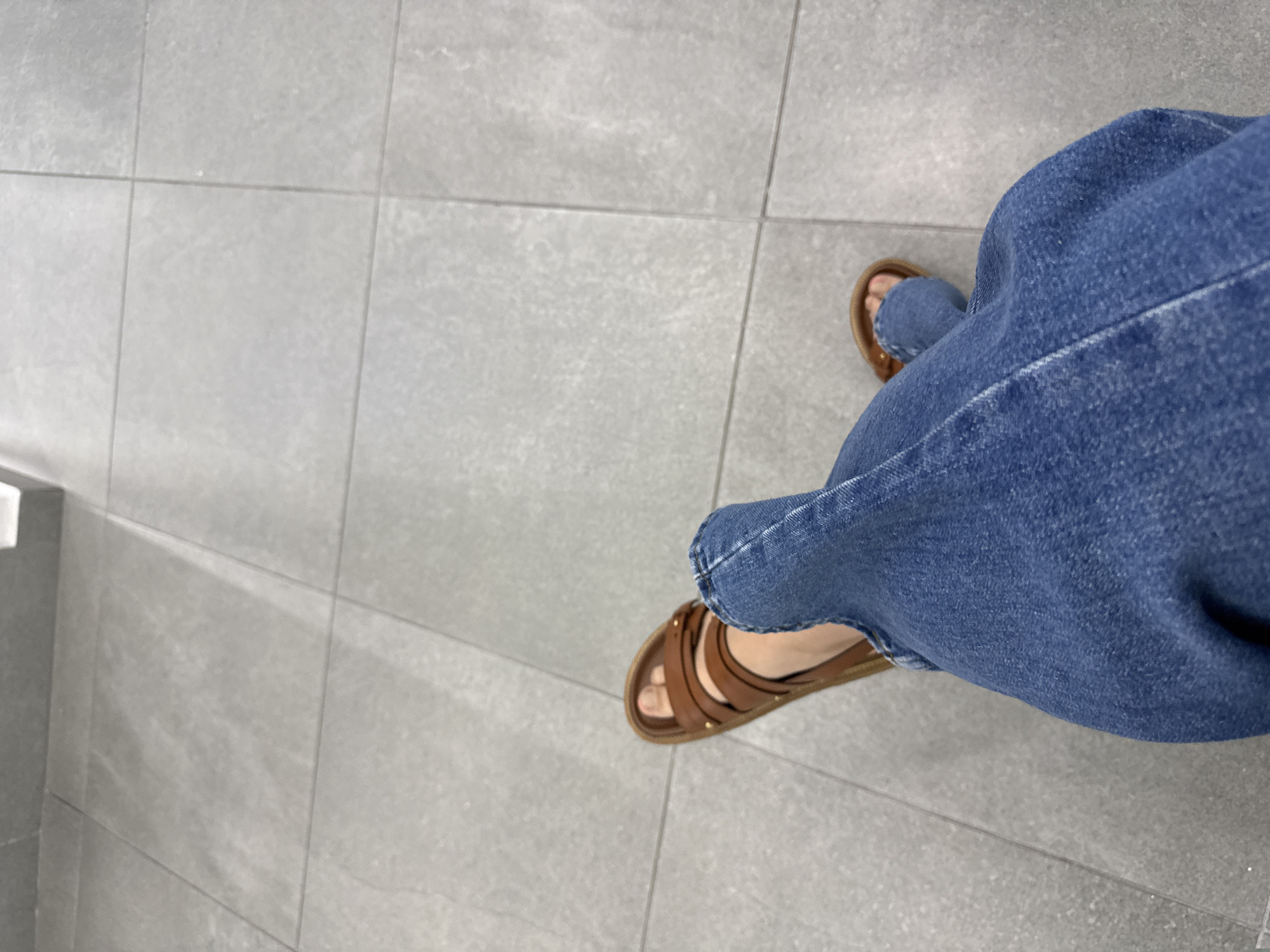 The image shows a person's legs from the knees down, clad in blue jeans and brown sandals. They are standing on a tiled floor with a light gray color. The floor is made up of square tiles with narrow grout lines. A partial view of a white object is visible on the left side of the image. The specific location in San Bernardino Tlaxcalancingo, Mexico cannot be determined without additional information.