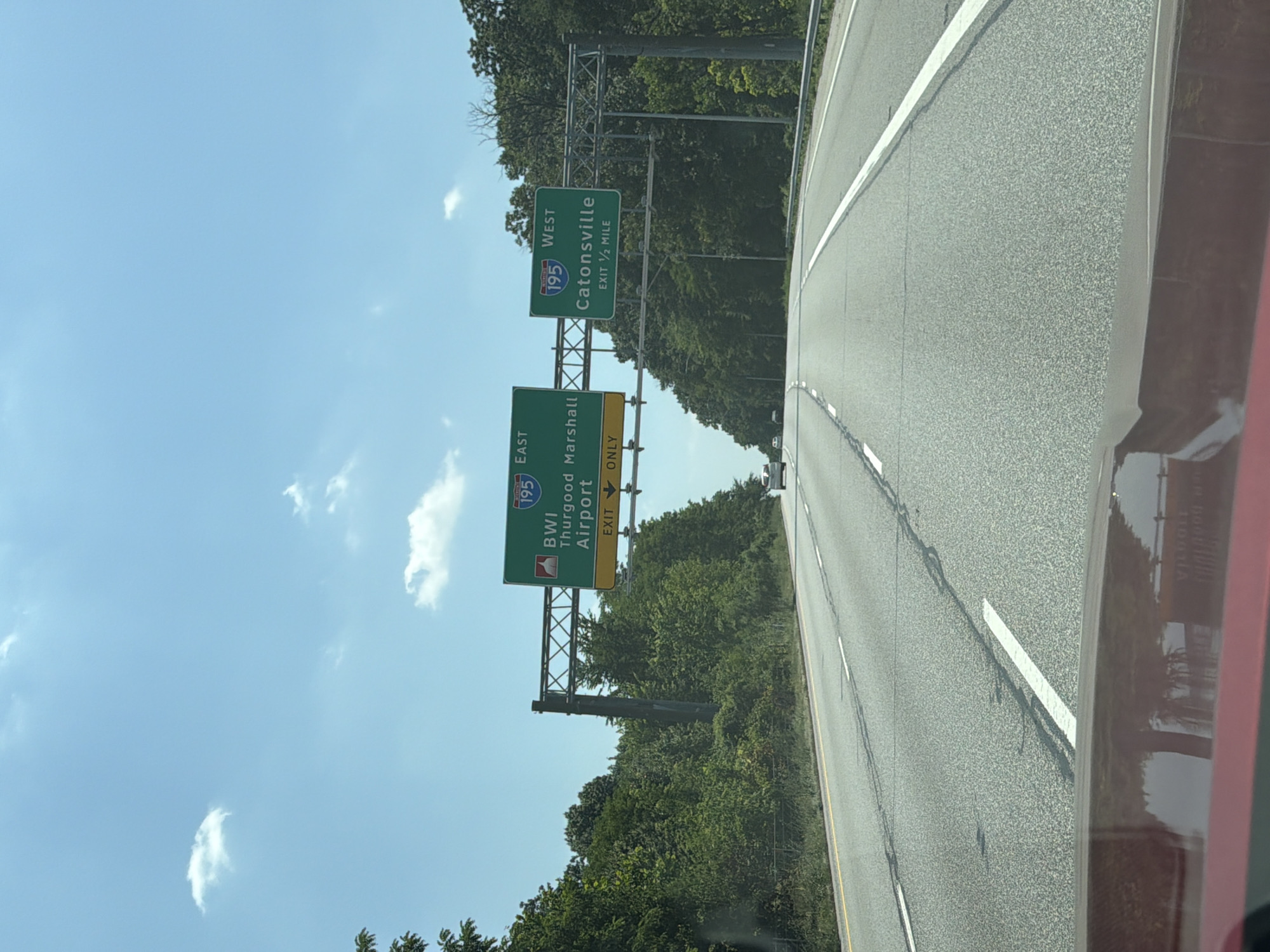 The image depicts a highway scene in Glen Burnie, United States of America, with overhead signage directing traffic. On the left, a green sign indicates "195 EAST BWI Thurgood Marshall Airport" with an "EXIT 12 ONLY" designation below. To the right, another green sign reads "195 WEST Catonsville" and states "EXIT 1/2 MILE". The road is a multi-lane highway with white lane markings. Trees and foliage line the sides of the highway. The sky is clear blue with a few scattered white clouds. The reflection of a red vehicle is visible in the foreground.