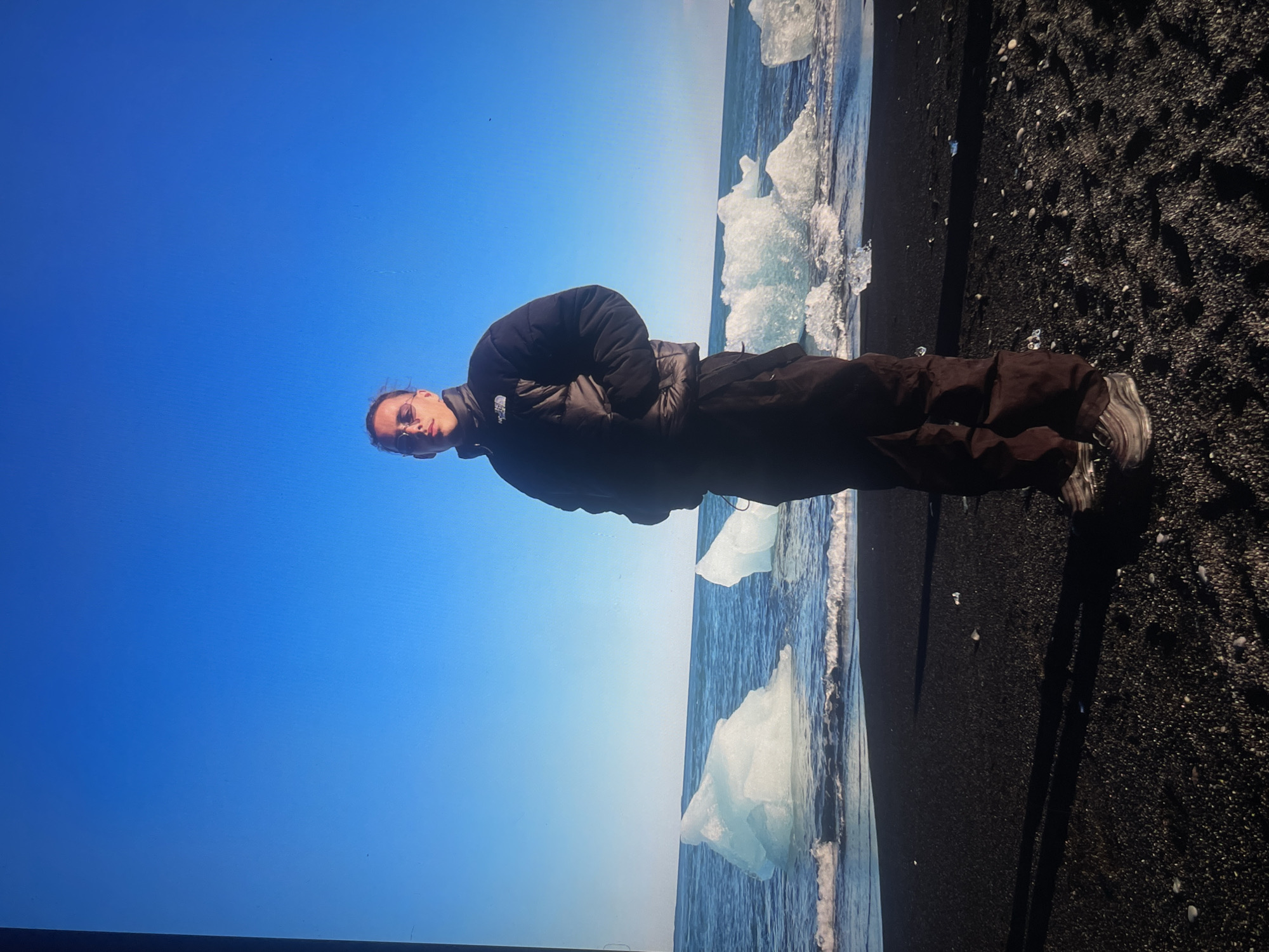 A person stands on a black sand beach in Iceland, next to the ocean with icebergs floating in the waves. The sky is clear and blue. The person wears a dark puffer jacket and dark pants.
