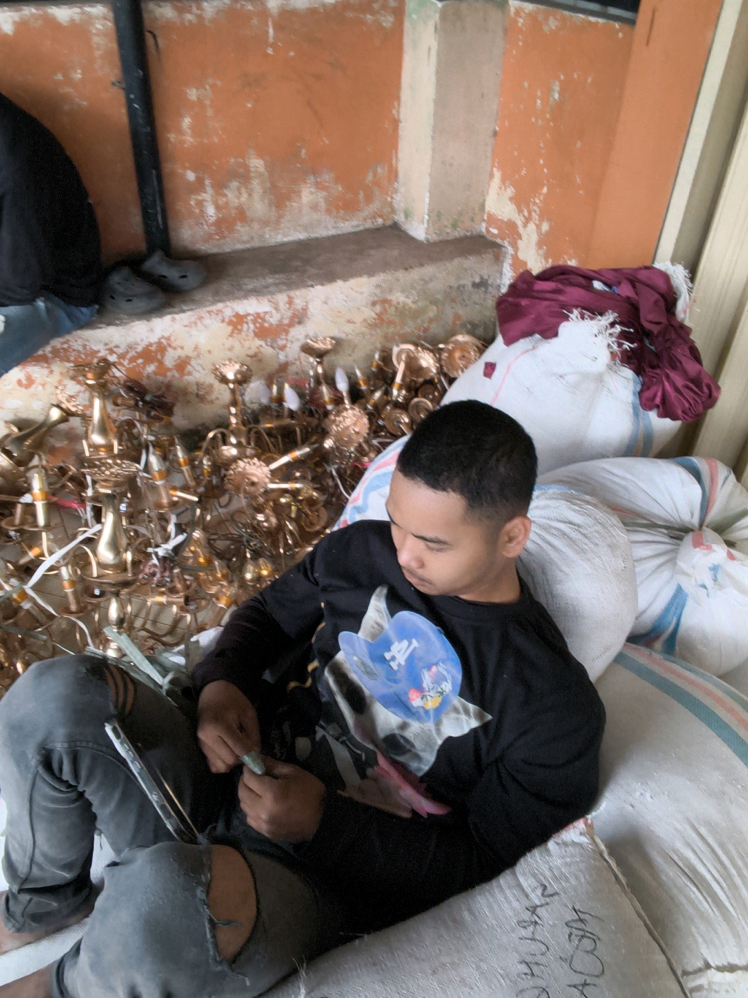 A young Indonesian male sits amidst a pile of golden chandeliers and large white sacks. He is wearing a black long-sleeved shirt with a graphic print and distressed grey jeans. His hands are occupied with something small and green, possibly a piece of candy or a small object. The background features an orange, textured wall, suggesting an outdoor or semi-outdoor industrial setting in Lindung Jaya, Indonesia. The overall scene appears to be a workspace or storage area.
