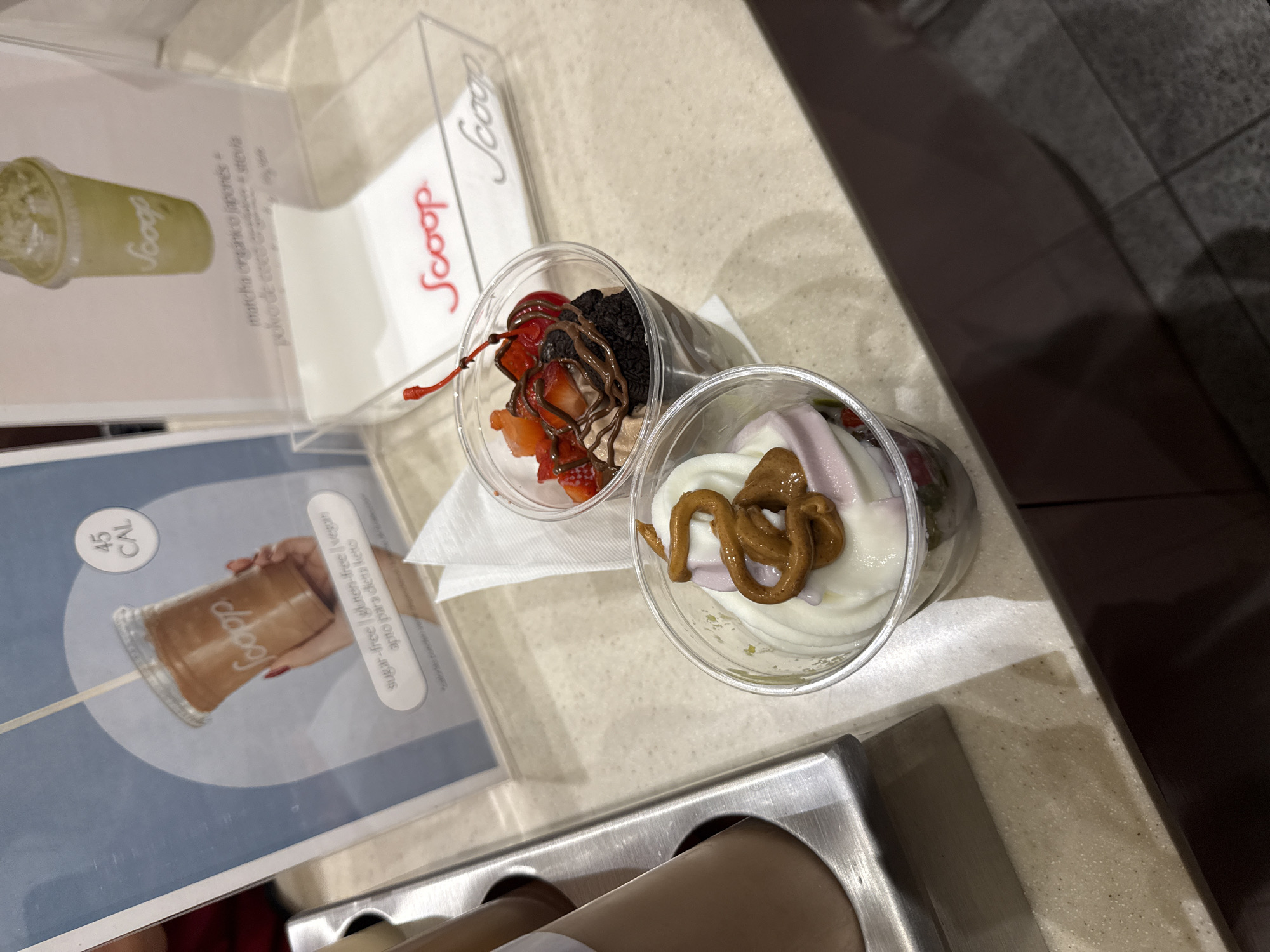 The image shows two cups of what appears to be frozen yogurt or a similar dessert, arranged on a tray or counter in what seems to be a business location with "Scoop" branding. One cup contains the frozen treat topped with what looks like caramel or a nut-based sauce. The other cup has toppings that include strawberries, chocolate sauce, and a cherry. Next to the cups are promotional displays with the "Scoop" logo, one for a matcha drink and the other for a sugar-free, gluten-free, vegan, and keto-friendly product. The image resolution does not allow me to determine the exact location within Mexico City, but the style of the branding and products indicates a modern dessert shop.
