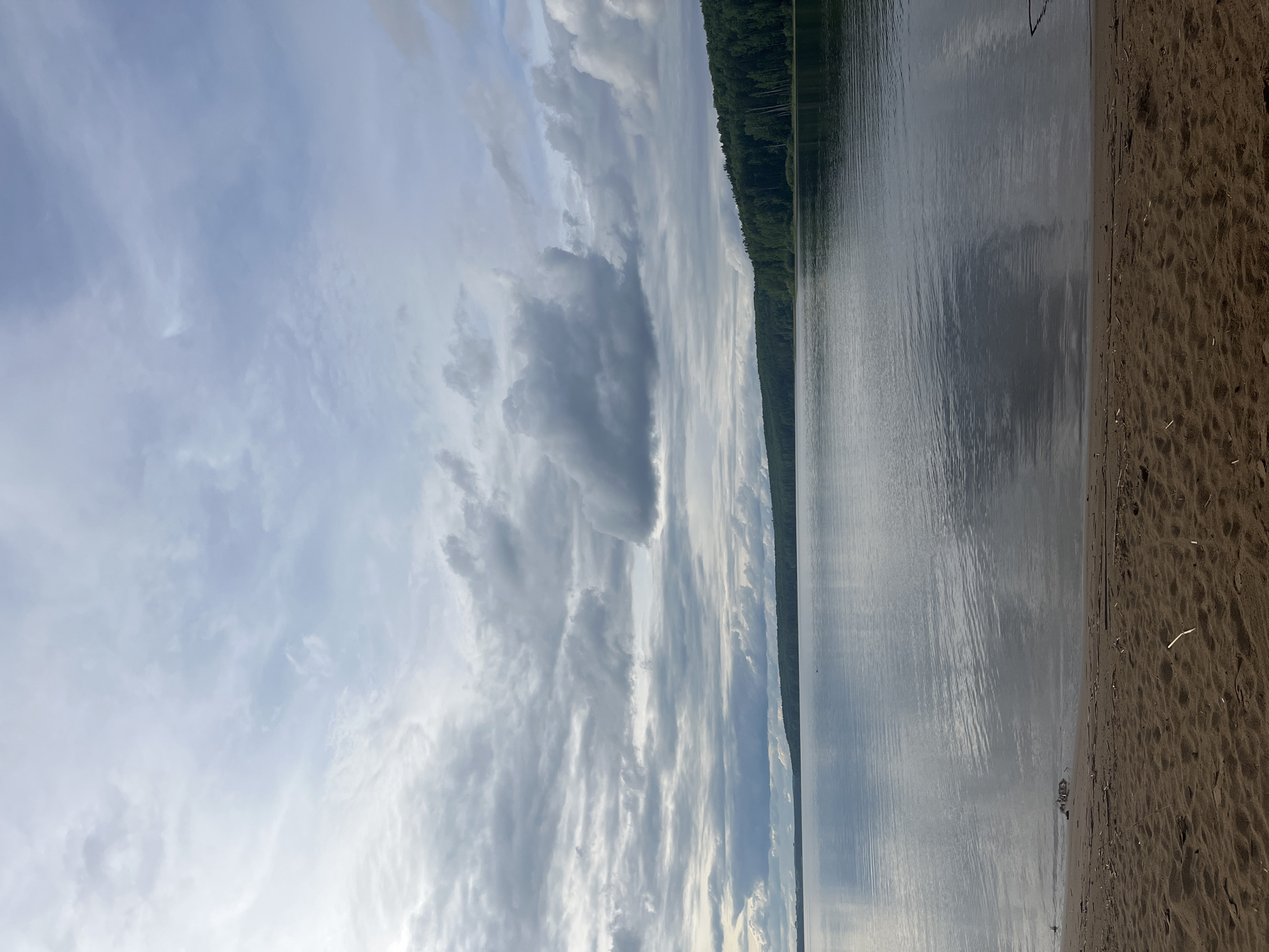 The image depicts a lakeside view under a cloudy sky. The sky is filled with layers of clouds, with variations in density and color. The lake's surface is relatively calm, reflecting the sky above. Along the shoreline, there is a strip of dark sand. A dense line of trees can be seen on the opposite shore. The location is Unknown, Unknown.