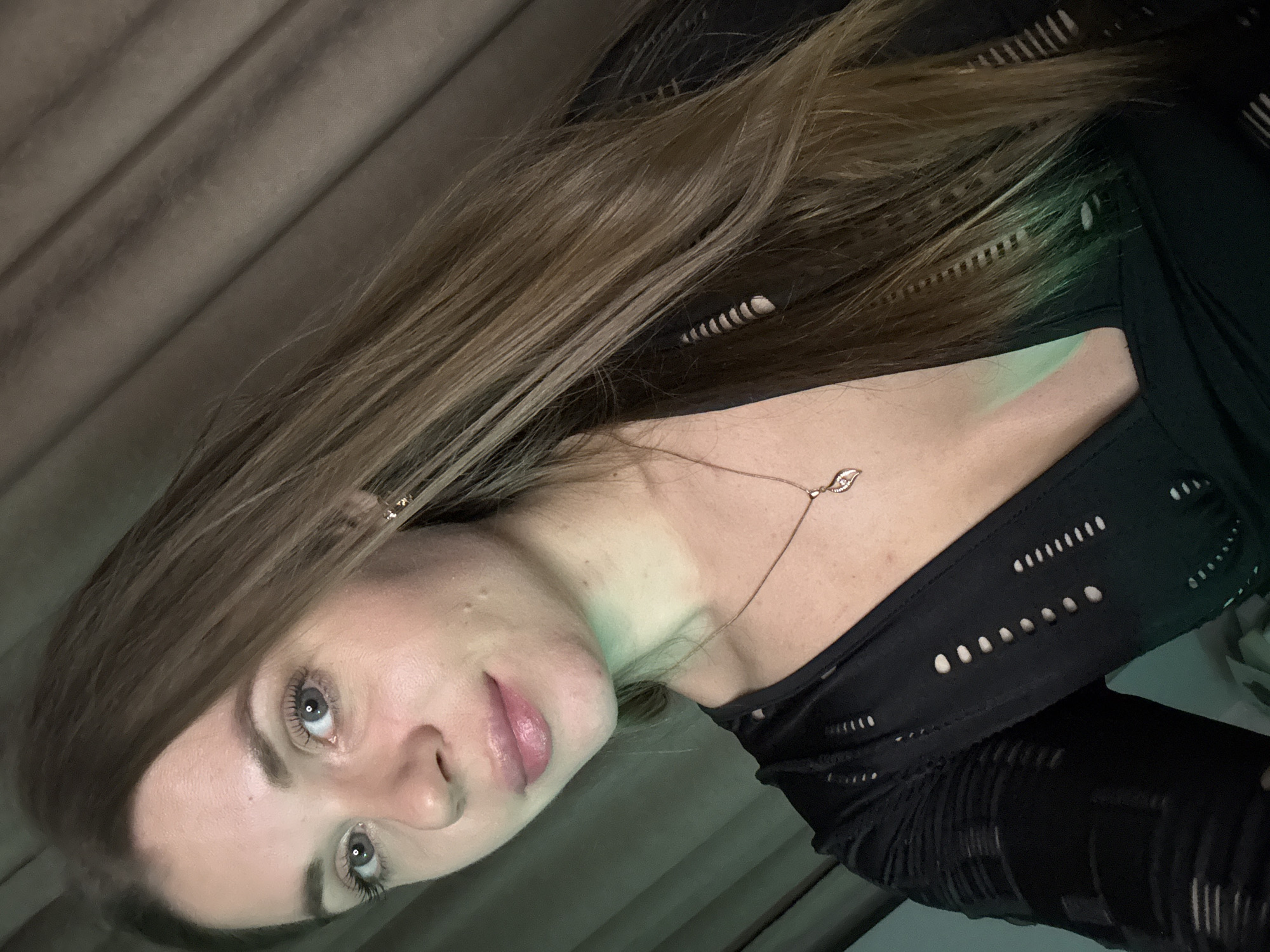 A close-up, angled portrait features a young woman with long, straight, light brown hair falling over one shoulder. She looks directly at the camera with a calm, slightly serious expression, revealing clear blue-grey eyes and full, pink-toned lips.
She is wearing a black top or dress with intricate cutout patterns visible on the shoulders, chest, and arms. A delicate rose gold-toned necklace with a small, leaf-shaped pendant, featuring a tiny gem, rests on her neck. Small, gold-colored stud earrings are partially visible.
A prominent green light casts a glow across her neck and upper chest, creating a striking contrast with her skin tone and dark clothing. The background appears to be an indoor setting with textured, possibly draped, walls in shades of brown or grey. The lighting suggests the image was taken during the evening or nighttime.
The woman's pose is a direct address to the camera. While the provided context indicates the location is Batumi, Georgia, there are no specific visual cues within the image to confirm this. No text is visible in the media file.