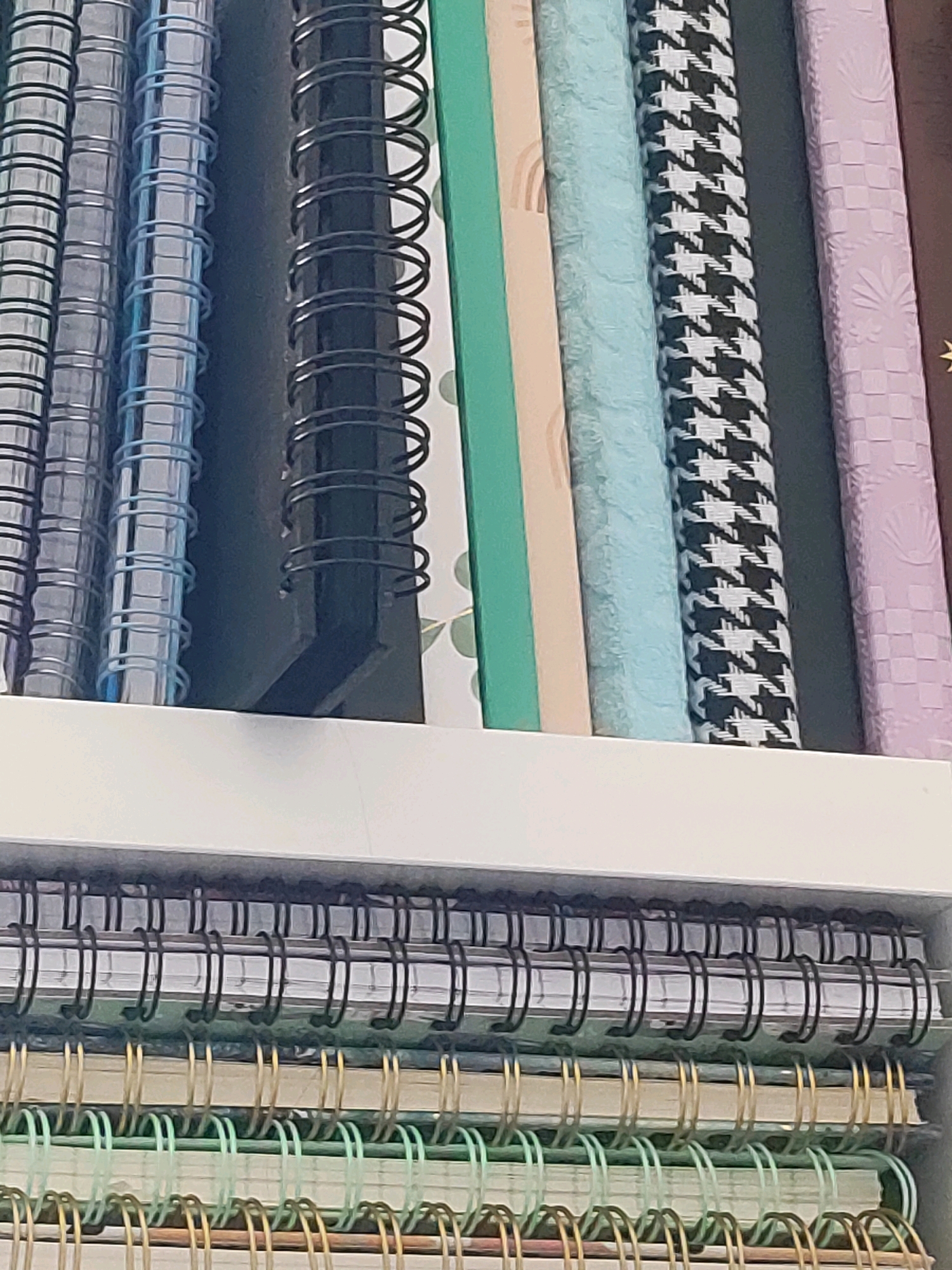 The image displays a collection of spiral-bound notebooks arranged vertically on a white shelf. The notebooks feature a variety of covers, including solid blue, black with a houndstooth pattern, and light purple with an embossed pattern. Some notebooks also have patterned covers with rainbows and a light green hue. Below the shelf, another row of notebooks is visible, with their spiral bindings in black, gold, and green. The notebooks are densely packed, with their spines facing the viewer. The scene appears to be an indoor setting, possibly a retail display or a personal collection. There is no indication of the city of Burgoberbach, Germany, in the image itself.