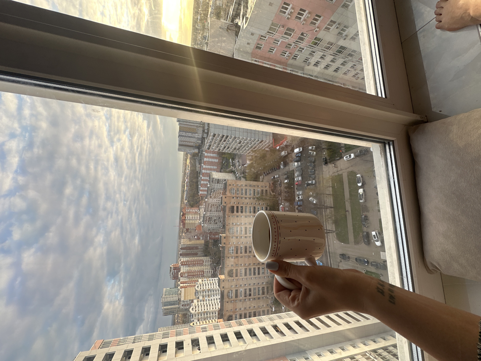 A high-angle shot taken from inside a dwelling shows a cityscape outside a window, with a person holding a mug. The window frame is visible, and the cityscape is dominated by multiple high-rise apartment buildings of varying colors and architectural styles. The sky is overcast with clouds. A row of parked cars and trees are seen between the buildings. The person is holding a light-colored mug with a pattern, and their arm has a tattoo on it. The photo was taken in Khimki, Russia.