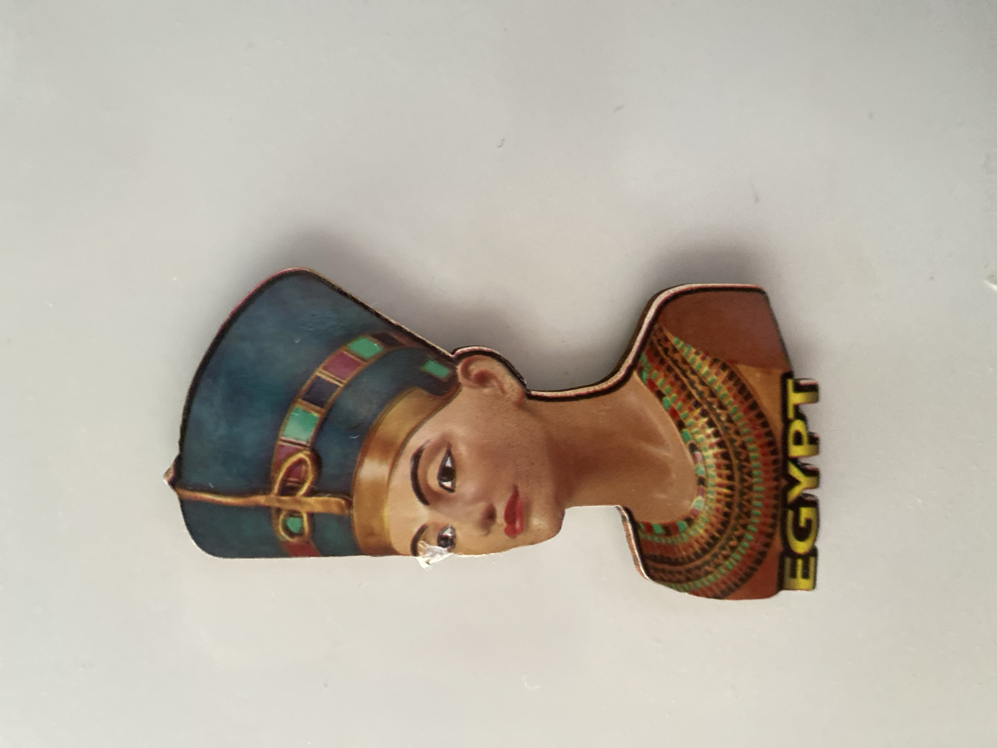 The image displays a souvenir magnet shaped like the bust of Nefertiti. The magnet features the queen wearing her distinctive blue headdress adorned with colorful bands of red, green, and gold. She is also depicted with a large, elaborate collar necklace in similar vibrant colors. The words "EGYPT" are printed in yellow text at the bottom of the magnet. The magnet is attached to a plain, light-colored wall, which serves as the background. There is no identifiable location in the city of Sokhumi, Abkhazia, visible in the image.