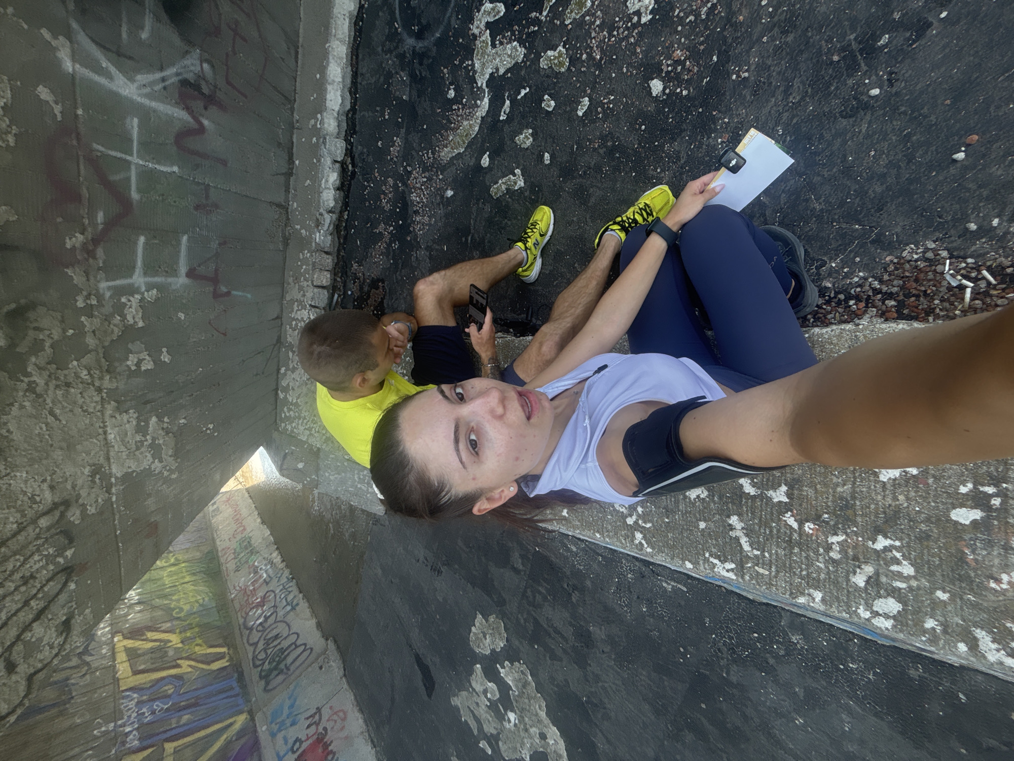 This is a selfie-style image taken from a low angle, showing two people sitting on a concrete ledge under an overpass. The background features a tunnel or underpass with graffiti on the walls and a small stream of water on the ground.
In the foreground, a woman is looking directly at the camera with an open mouth. She has her hair pulled back in a ponytail and is wearing a white sleeveless top, dark blue leggings, and a black armband. She is holding a white piece of paper with a small black clip attached to it.
Behind her and to the right, a man is sitting with his legs extended. He is looking down at his phone and is wearing a bright yellow t-shirt and dark shorts. He has bright yellow athletic shoes on.
The environment appears to be urban and somewhat neglected, with rough concrete surfaces and evidence of graffiti. The presence of the water and the graffiti suggests it might be a drainage area or an underpass commonly used for recreation or as a canvas for street art. The overall lighting suggests it is daytime. The exact location in Varna, Bulgaria, is not specified in the image itself, but the graffiti style and general ambiance are consistent with urban underpasses found in various cities.