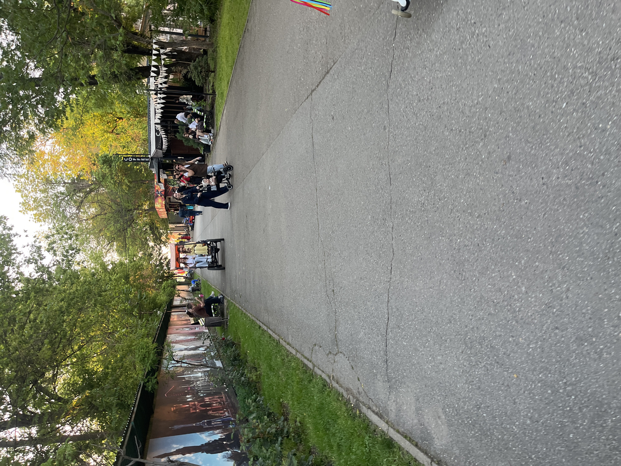 The image shows a pedestrian walkway in a park in Krasnodar, Russia. On the left, there are trees, benches with people sitting on them, and a fence with images. On the right is a grassy area and the paved walkway. Several people are riding quadricycles along the path. There are buildings and a food vendor in the background.