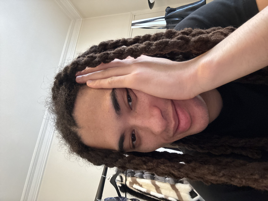 The image displays a close-up portrait of an individual with dark skin and long, dark brown dreadlocks, looking downward and to their left with a subtle smile. Their right hand rests against the right side of their face, fingers extended upwards, with the palm open. The person is wearing a black t-shirt. The background to the left reveals a clothing rack holding various garments, prominently featuring a patterned brown, beige, and black fur or faux fur coat. To the right, a light-colored wall is visible, along with a black bag with white vertical stripes and another dark, indistinct object. White crown molding runs along the top of the wall, indicating an indoor setting. There are no visual cues within the image to ascertain a specific location within the city of Paris, France.