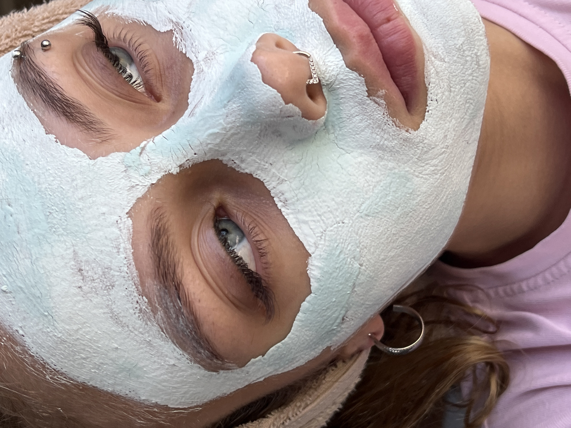A close-up, top-down, eye-level shot shows the face of a person wearing a pale blue face mask that covers most of their skin, obscuring their forehead, cheeks, and nose. Their left eye, with long eyelashes and blue-green iris, is clearly visible, looking towards the left side of the frame. A silver eyebrow piercing is visible above the eye. The person has a small, silver hoop earring in their left earlobe and a decorative silver nose ring on their right nostril. Their lips are partially visible and are tinted pink. They are wearing a light pink t-shirt and a beige towel is draped around their head. The scene is indoor and there are no visible elements that indicate a specific location in Fokino, Russia.