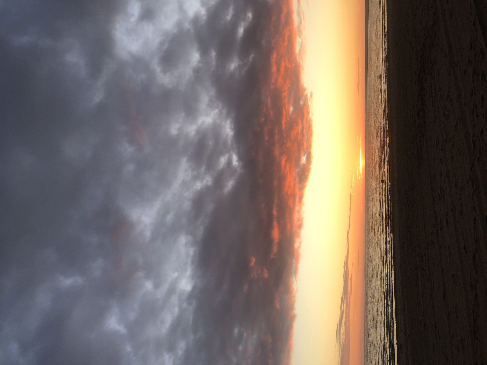 This is a vertical, eye-level, full shot of a sunset over a beach and the Gulf of Finland in Sestroretsk, Russia. The foreground is a dark, sandy beach with some faint footprints. The middle ground is the calm, dark water of the gulf, with subtle ripples reflecting the sky. A buoys is visible in the water towards the right. In the distance, the horizon is visible, with a faint landmass. The sky is dominated by dramatic clouds. The lower part of the sky is a vibrant gradient of yellow and orange, indicating the setting sun, which is a bright, horizontal line just above the water. Higher up, the clouds are dark gray, with touches of orange and pink where they catch the last rays of the sun. The overall mood is serene and awe-inspiring.