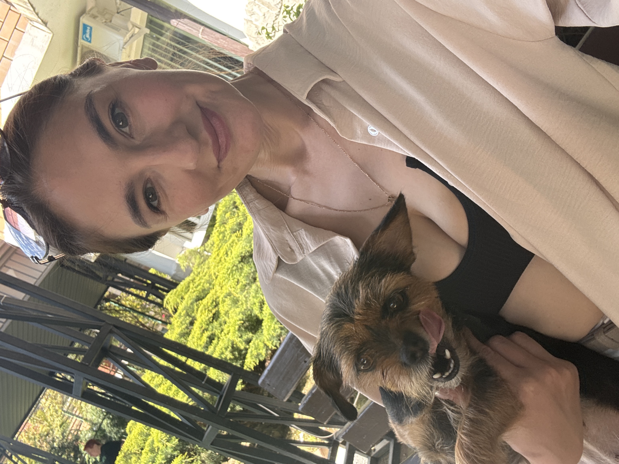 A woman holds a small brown and black terrier-type dog, appearing to lick its face. The woman has brown hair pulled back, sunglasses resting on her head, and wears a light beige shirt over a black top. She is wearing a thin gold necklace. The background shows a metal structure, green trees, and a building with an air conditioning unit. Based on the style of buildings, vegetation, and metal structure, the location is consistent with outdoor cafes or patios in Sochi, Russia.