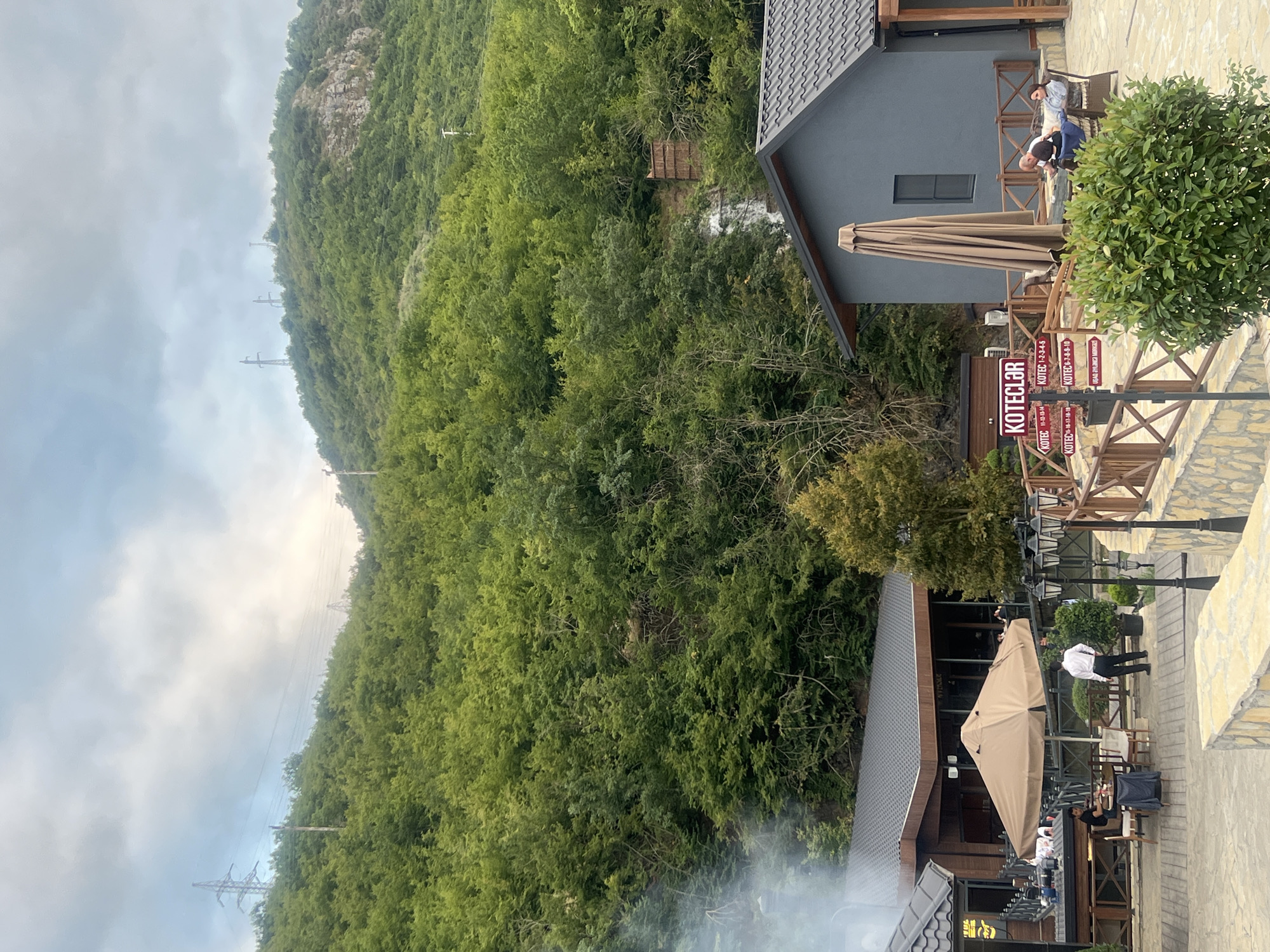 In the city of Khankendi, Azerbaijan, a multi-story restaurant complex is situated at the base of a lush, green hillside covered in trees. The sky is overcast with scattered clouds. In the foreground, to the left, is a section of the restaurant with dark roofing and wooden accents. An outdoor seating area with tables and chairs is visible under a large, beige umbrella. A waiter in a white shirt and black pants stands near a table. To the right, another building with dark gray walls and a dark tiled roof is partially visible. Two people are seated on chairs outside this building, conversing. A thick green bush is in front of them. Several directional signs are posted on a wooden post, with text in an unfamiliar script. The environment is natural and mountainous, with power lines visible against the sky on the hillside.