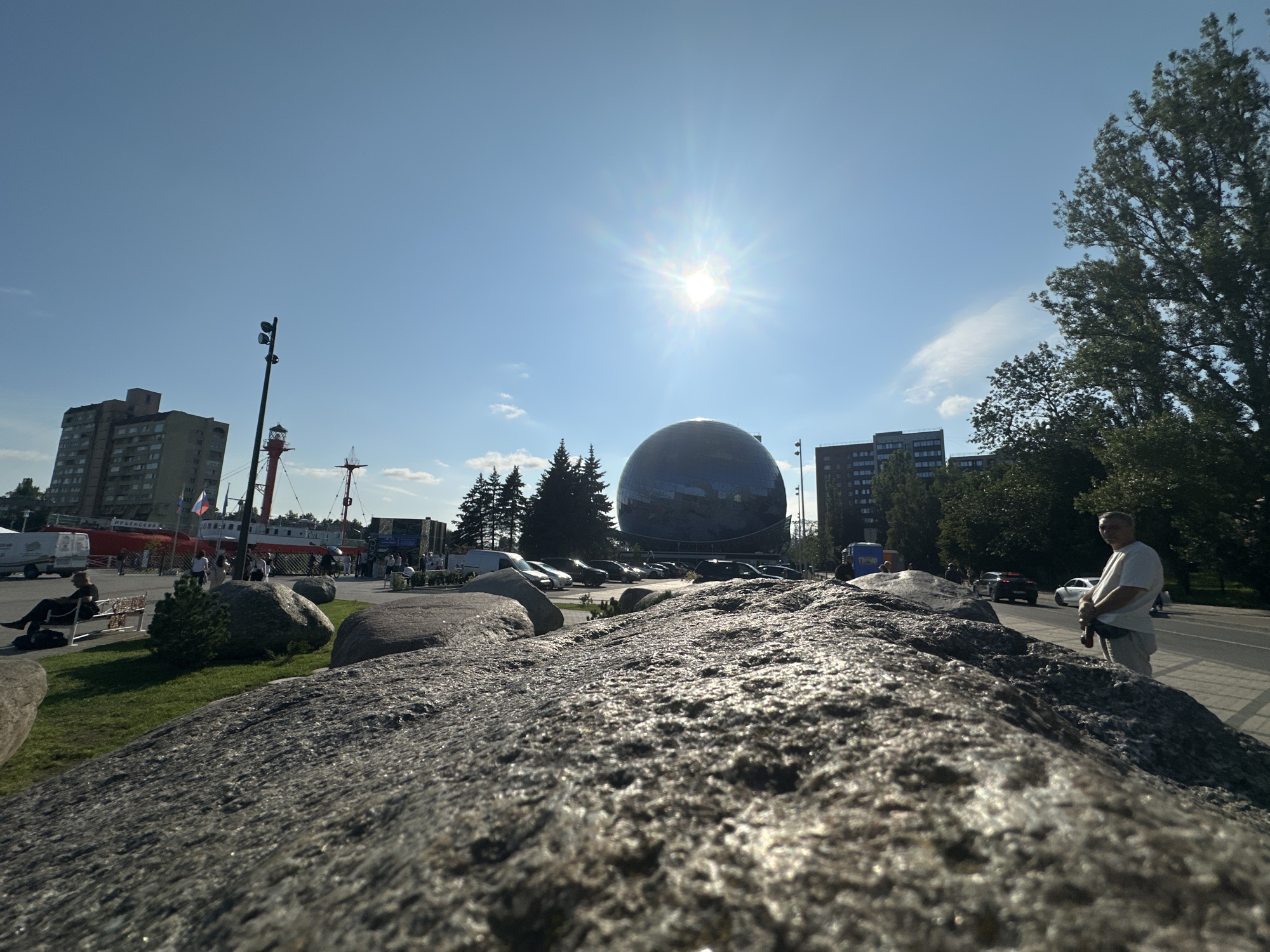 The image depicts an outdoor scene on a sunny day in Kaliningrad, Russia. In the foreground, large granite boulders dominate the lower portion of the frame, with one prominently placed in the foreground, its textured surface catching the sunlight. Beyond the boulders, a green grassy area is visible with a few people sitting on a bench.
In the midground, a large, reflective spherical building, likely the Kaliningrad Planetarium, stands out against the clear blue sky. Several evergreen trees are positioned in front of the planetarium. A red and white ship is docked nearby, with people milling around on the quay. A lighthouse structure is also present.
In the background, there are multi-story apartment buildings and other structures, characteristic of an urban environment. A road runs alongside the scene, with cars and a few pedestrians visible. One man, wearing a white t-shirt and light-colored pants, stands on the sidewalk, looking towards the camera. The sun is high in the sky, casting bright light and creating sun flares. The overall impression is of a public space in a city, likely a waterfront area, on a bright summer day.