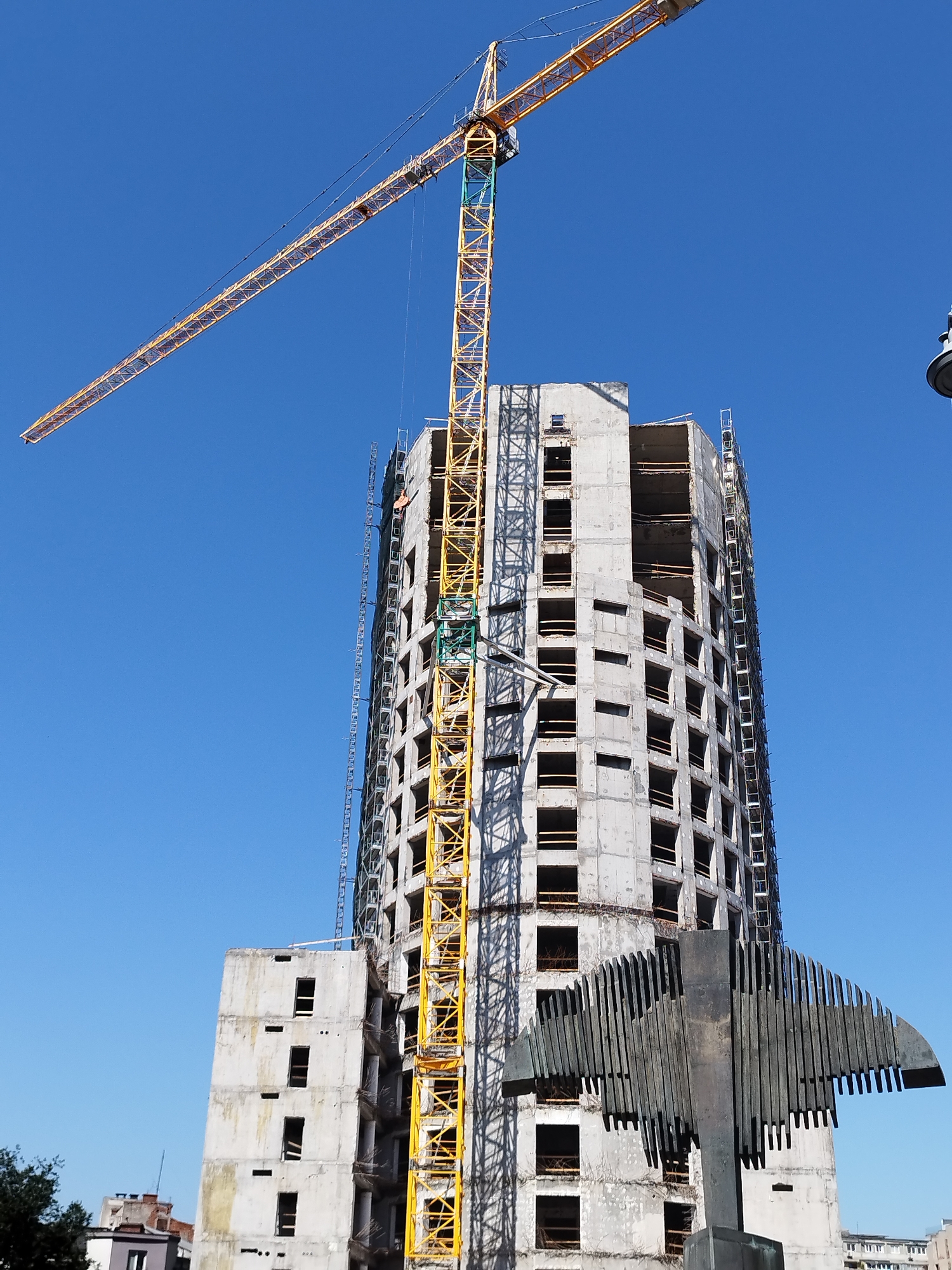The image depicts a construction site in Bucharest, Romania, featuring a partially constructed skyscraper and a large yellow tower crane. The skyscraper is concrete and has an unfinished facade with numerous open window frames. The tower crane is positioned alongside the building, with its long boom extending horizontally across the frame against a clear blue sky. In the foreground, a modern sculpture made of dark metal with vertical slats and a curved wing-like structure is visible. The scene suggests ongoing construction work on a significant urban development project.
