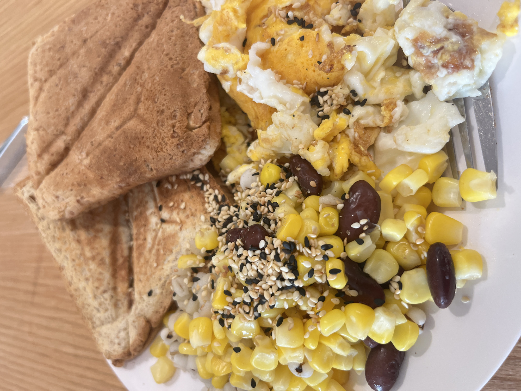The image displays a close-up, overhead view of a prepared meal served on a white plate, which rests on a light-colored wooden surface. The meal appears to be a breakfast or brunch offering.
The main subjects of the image are various food items. On the left side of the plate, two pieces of lightly toasted bread, possibly whole-grain, are folded or arranged. Adjacent to the toast, a generous serving of scrambled eggs occupies the center of the plate, characterized by both yellow yolk and white albumen portions. These eggs are sprinkled with a mix of white and black sesame seeds.
Below the scrambled eggs, a colorful medley consists primarily of bright yellow corn kernels mixed with dark, reddish-brown beans, which resemble kidney beans. This corn and bean mixture is also garnished with a scattering of white and black sesame seeds. A silver fork is partially visible on the right side of the plate, suggesting the meal is ready for consumption.
The scene is a dining setting, likely indoors, given the soft, even lighting. There are no people visible in the image. The food is presented neatly, highlighting its textures and colors. No visible text is present in the image. While the location context is provided as Pattaya City, Thailand, there are no specific visual cues within the image itself to confirm this geographic location.