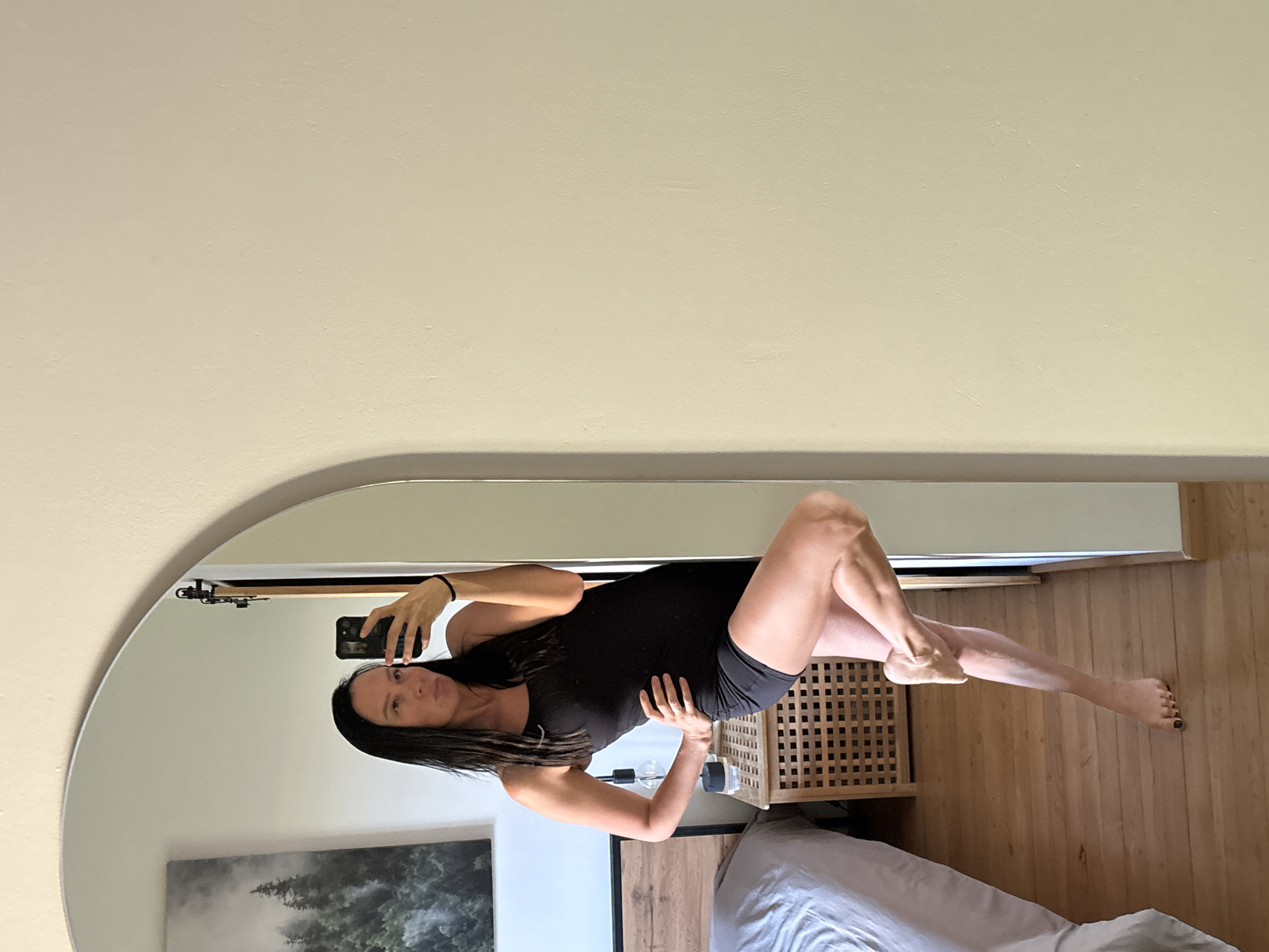 A woman is taking a selfie in a mirror. She has long dark hair and is wearing a black tank top and black shorts. She is standing with one leg bent and crossed over the other. Her left hand is on her hip. The mirror is in a bedroom. In the reflection, a bed with grey bedding is visible on the left side of the image, along with a wooden nightstand. On the wall behind the bed is a framed picture of a forest. The room has light-colored walls and a wooden floor.