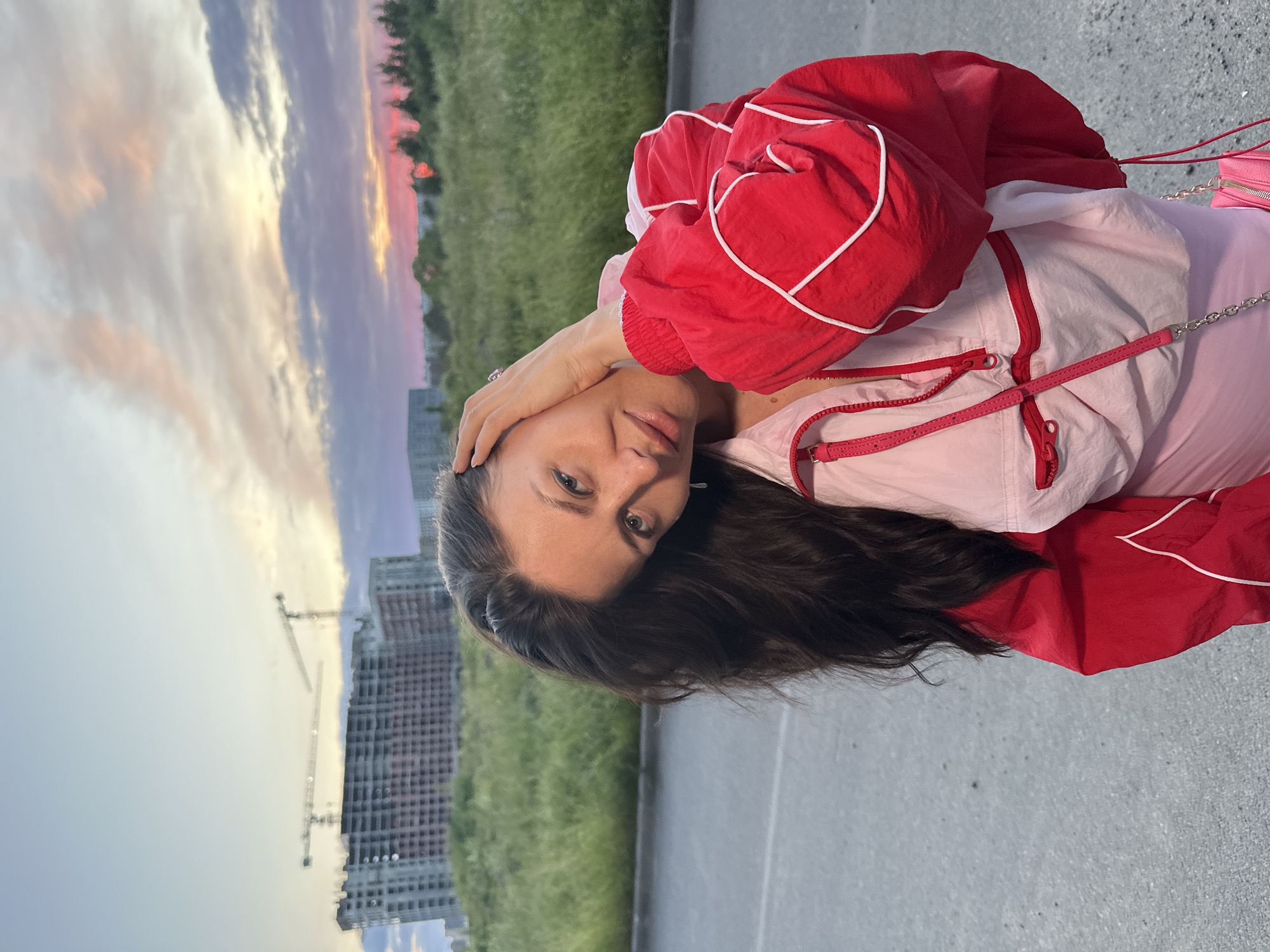 In Murino, Russia, a woman with long, dark hair poses against a backdrop of a construction site and a vibrant sunset. She is wearing a bright red and pink jacket, and a pink purse with a chain strap is visible. Her left hand is placed on her head, and her right hand is gently touching her neck. The sky displays a palette of pink, purple, and orange hues, indicative of sunset. The foreground consists of an asphalt surface, with a grassy area and an unfinished building with cranes in the mid-ground.