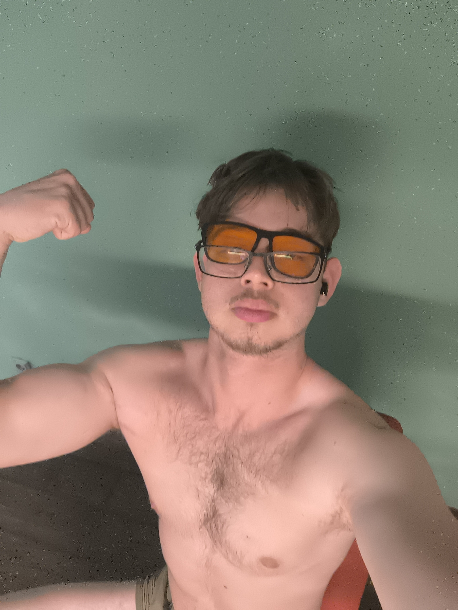 A young man is the central subject of this indoor, waist-up shot. He is shirtless, displaying a light covering of body hair on his chest and arms. He has short, messy brown hair and light facial hair in the form of a goatee or stubble. His lips are slightly parted, and he has a neutral to slightly serious expression as he looks directly at the camera.
He is wearing dark-rimmed glasses with distinct orange-tinted lenses, covering his eyes. A black earbud is visible in his right ear. The man is posing by flexing his left bicep, with his fist clenched. His right arm is relaxed at his side.
The background is a plain, textured greenish-blue wall. There appears to be some shadow on the wall to the right of the man's head and shoulder. The lower left portion of the image shows a darker, indistinct area, possibly a floor or furniture. A hint of a reddish-orange object, possibly a chair, is visible behind his right shoulder and arm. The lighting appears to be artificial.
There is no visible text in the image. The scene takes place in Sofia, Bulgaria.