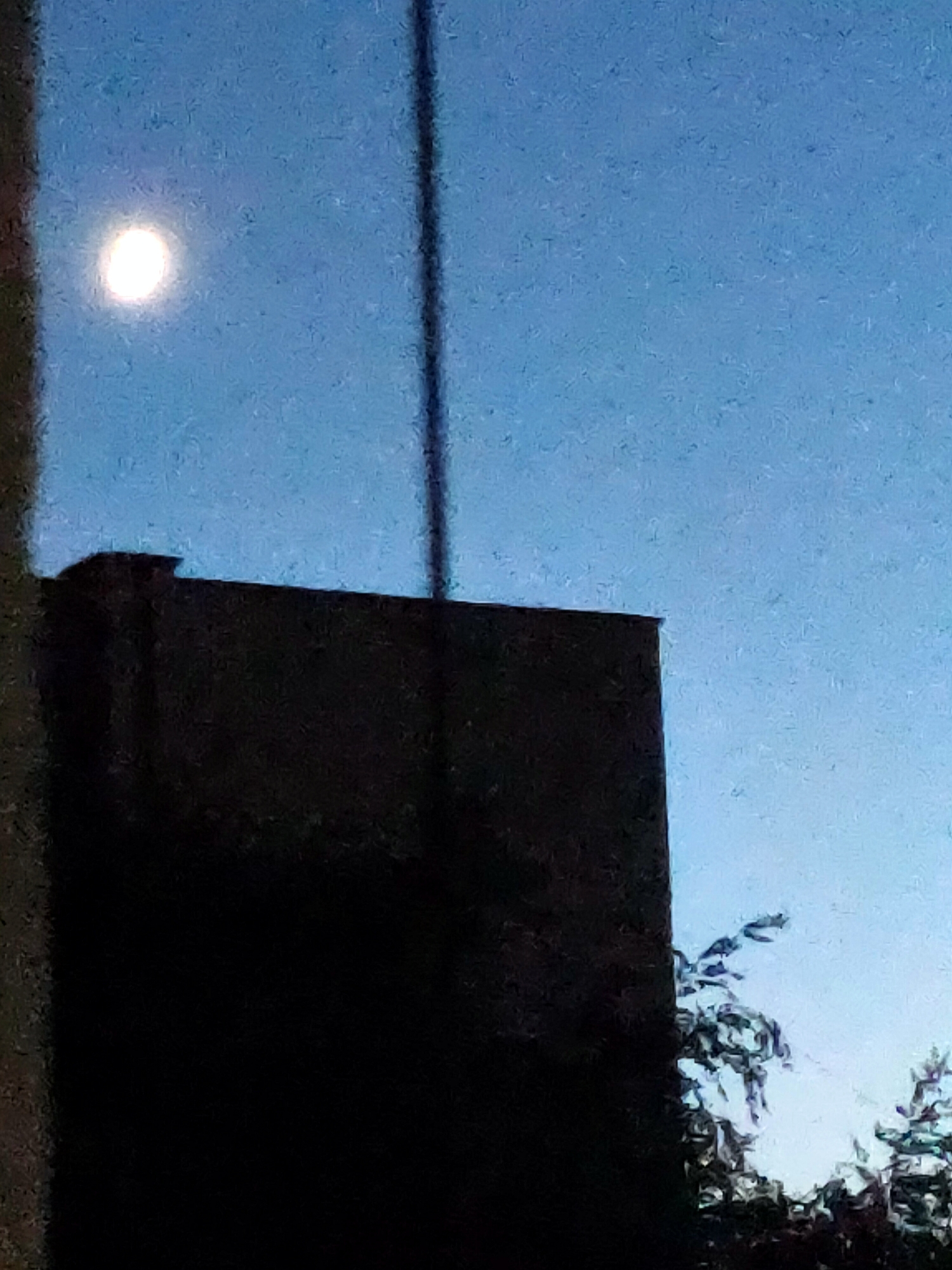 The image captures a twilight scene in Nosivka, Ukraine, featuring a bright, full moon positioned in the upper left quadrant of a clear blue sky. A dark, silhouetted building dominates the lower portion of the frame, with a visible chimney on its left side. A thin, dark vertical pole or antenna bisects the sky, running from the top to near the building's roofline. To the right of the building, the upper branches of trees are visible against the sky, their leaves appearing dark and indistinct. The overall impression is one of a quiet, nocturnal urban landscape under the illumination of the moon.