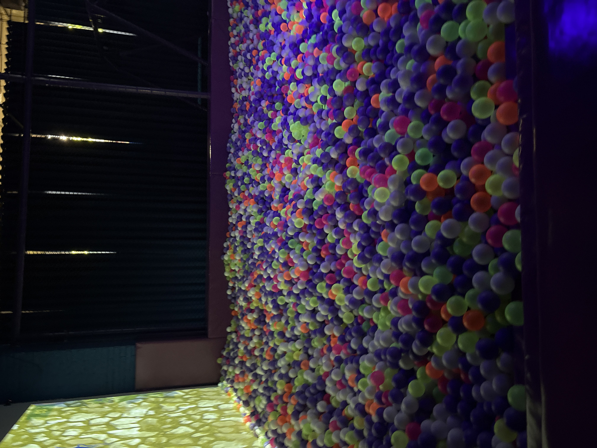 The image captures a room with a striking feature wall covered in colorful balls – predominantly shades of purple, green, orange, and pink. The wall is densely packed with these balls, creating a vibrant and textured surface. To the left, a darker area reveals a corrugated metal wall, possibly part of the building's structure, with some lighting reflecting off its surface. The foreground shows a section of floor with a light-colored, possibly projected, design resembling a natural scene. The lighting in the room casts strong shadows and highlights the colorful balls, emphasizing their presence. The location information, Izhevsk, Russia, suggests this might be an entertainment venue or a space designed for interactive experiences.