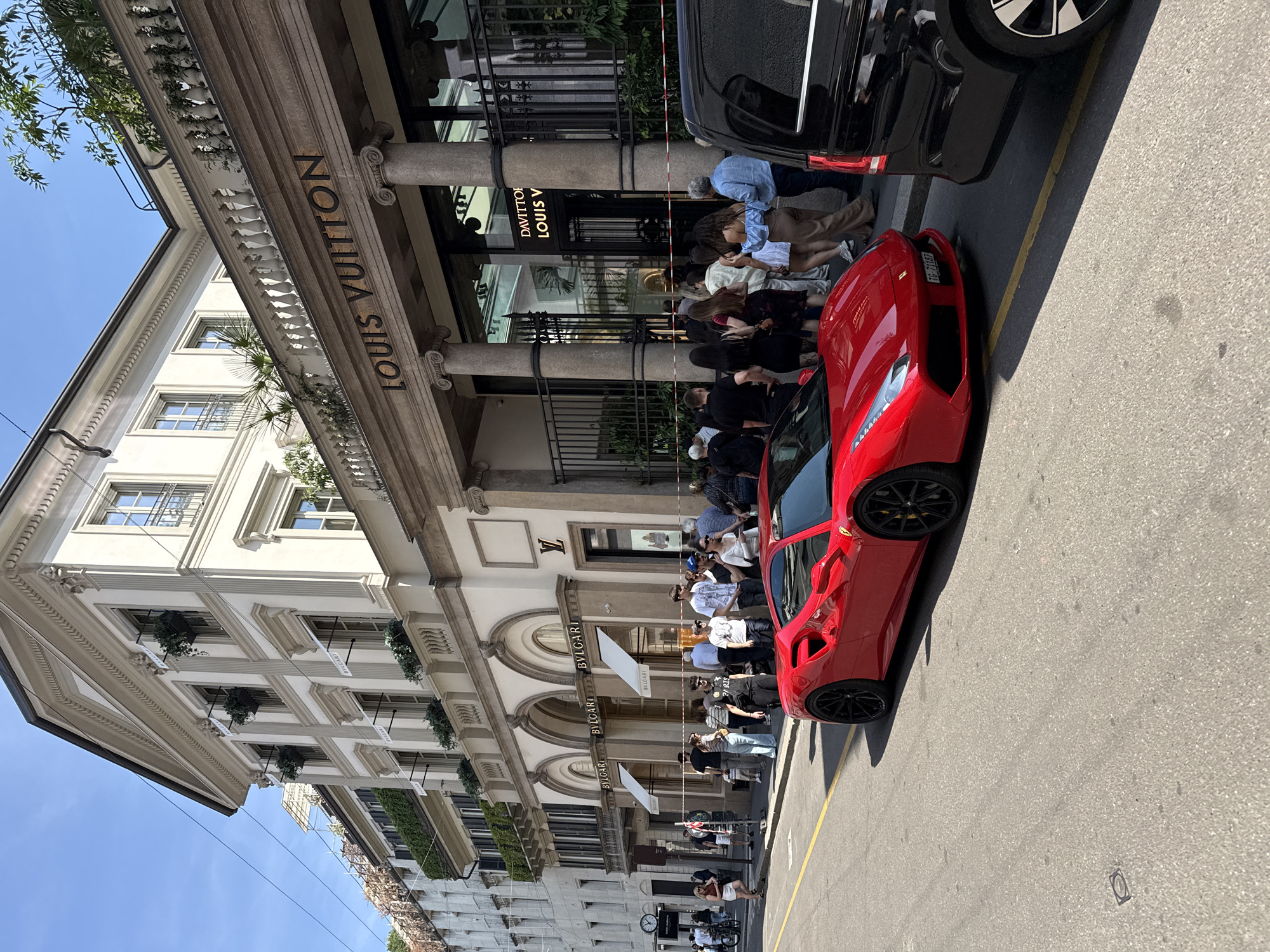 The image captures a scene on a street in Milan, Italy. Prominent luxury brand stores of Louis Vuitton and Bulgari occupy the buildings on the left side. A line of people stands in front of the stores. A red sports car is parked on the right side of the street, close to a black car. The sky is clear.