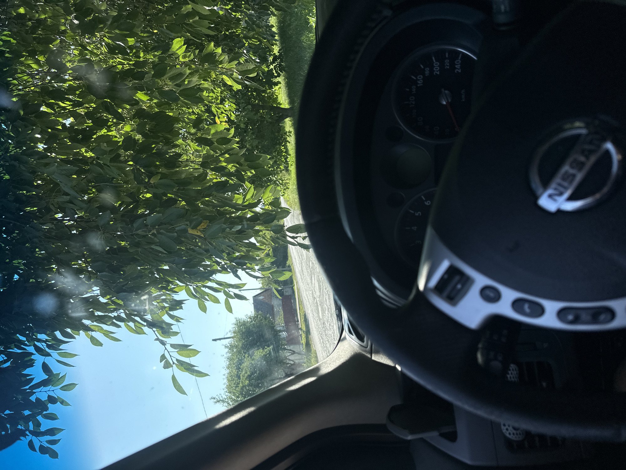 The image is a first-person perspective from inside a Nissan vehicle, looking out through the windshield. Sunlight filters through the green leaves of a tree that overhangs the road. In the foreground, the steering wheel and part of the dashboard are visible. The dashboard includes a speedometer with markings up to 240 km/h and the Nissan logo on the steering wheel. Outside the car, a rural road is visible, lined with trees and a house with a red roof. The sky is clear and blue. The scene is set in the village of Shatava, Ukraine.