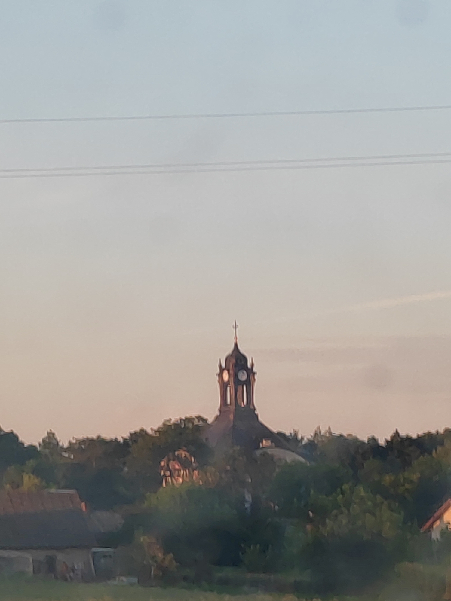 The image depicts a landscape view of the village of Volchinsk, Volchinski Selsoviet, Kamenets District, Brest Region, Belarus. In the center of the frame, a church tower with a clock and a cross stands tall above a dense canopy of green trees. To the left of the church, a portion of a building with a dark, sloped roof is visible, and to the right, another building with an orange roof is partially seen. The sky above is a soft gradient of pale blue and peach, suggesting either sunrise or sunset. Two thin, horizontal lines, likely power lines, traverse the upper portion of the sky. The overall impression is one of a serene rural setting dominated by the prominent church structure.