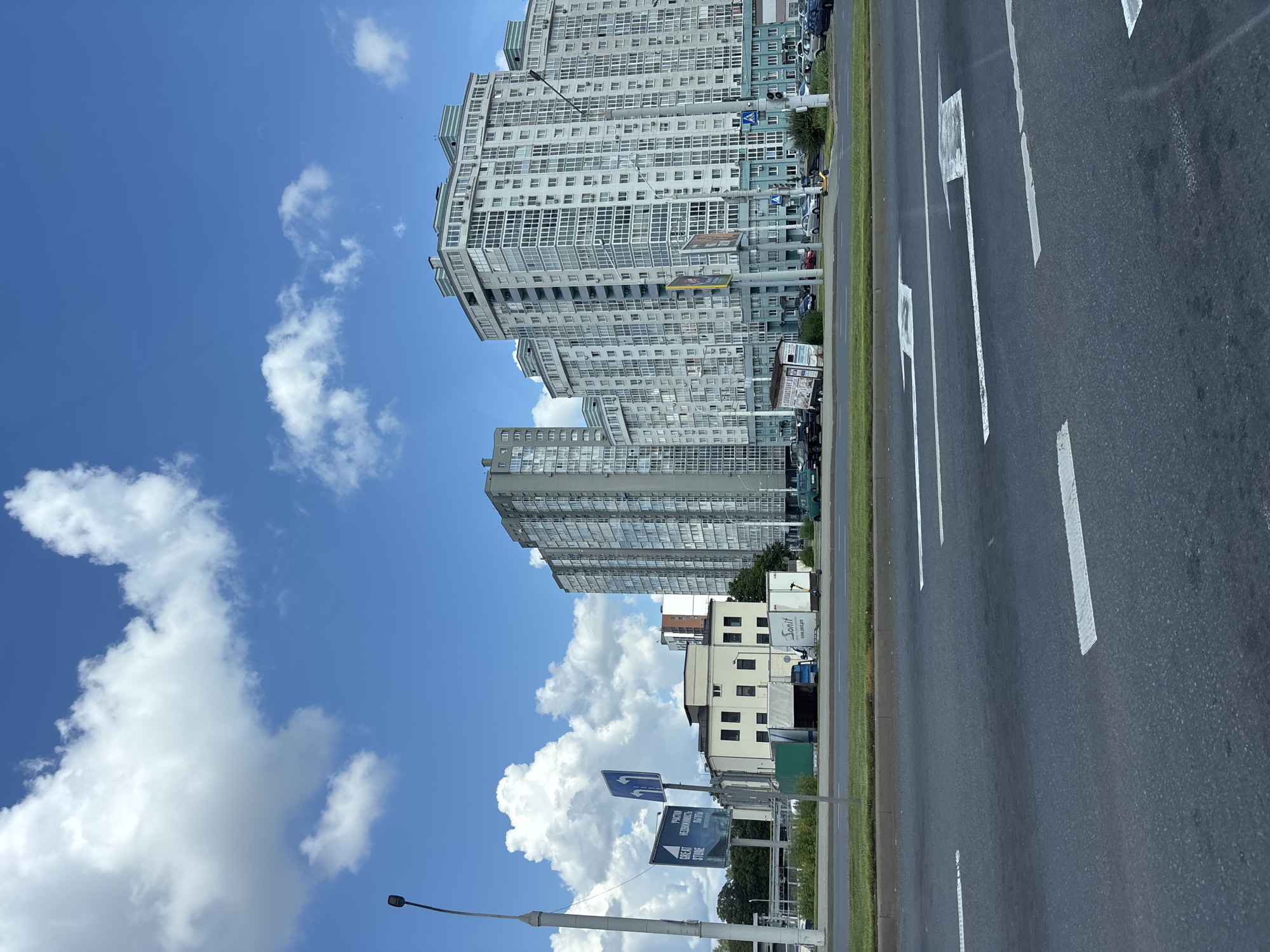 The image shows a street scene in Minsk, Belarus, on a partly cloudy day. In the foreground, a multi-lane asphalt road is visible with white directional arrows painted on the lanes. Beyond the road, there is a green median strip and then a sidewalk area.
On the sidewalk, a variety of structures and vehicles are present. To the left, a tall lamppost stands. Next to it are traffic signs, one with a blue directional arrow pointing left and another with blue symbols. Further to the right, there is a low, light-colored building with several dark windows. Adjacent to this building, there are temporary structures or booths, including a white trailer with "Sonit" and "www.sonit.pro" written on it. A yellow electric scooter is parked near this trailer.
Dominating the background are several large, modern apartment buildings. The building directly behind the low light-colored structure is a tall, grey, rectangular building with many windows. To its right, and slightly further back, is a much larger, multi-story apartment complex with a light-colored facade and numerous windows, many of which are floor-to-ceiling. This complex has a distinctive light blue trim along its upper floors and balconies. Other apartment buildings of varying heights and styles are visible further in the background. Several cars are parked and driving on the road in front of these buildings. The sky above is a vibrant blue, with large, fluffy white cumulus clouds scattered across it.