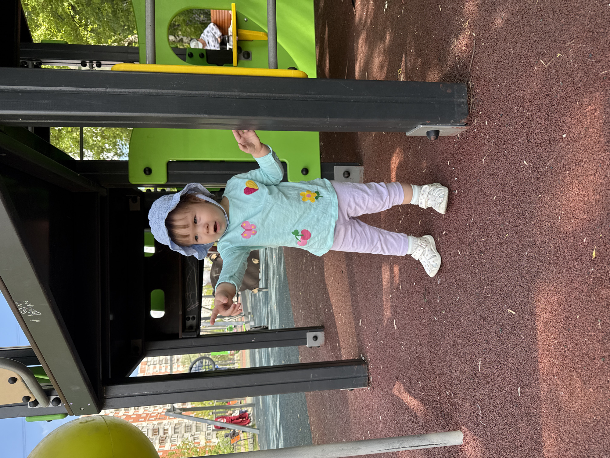 A young child in a blue hat, light blue long-sleeved shirt with flower and fruit appliques, and lavender pants stands on a red surface next to a playground structure. The structure is painted in black, green, and yellow. The child points to the left. The scene appears to be a playground. In the background, an apartment building is visible, indicating an urban setting. The location is Moscow, Russia.