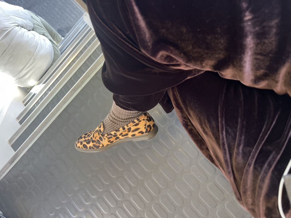 The image depicts an interior scene in Torcy, France, viewed from a slightly elevated, first-person perspective. In the foreground, the lower left leg of a person is visible, clad in dark brown, possibly velvet or plush fabric, trousers. The foot wears a leopard-print loafer with a dark sole, over a light brown ribbed sock. The floor beneath is dark grey, featuring a repetitive pattern of raised, oval-shaped textures. In the background, a set of light-colored stairs ascends towards the upper right, each step equipped with metallic or silver-toned anti-slip strips. Seated on one of the upper steps is a second individual, partially visible from the lower back. This person wears a light-colored, likely white or light grey, padded jacket and light green or khaki-colored trousers. No direct actions or interactions are depicted.