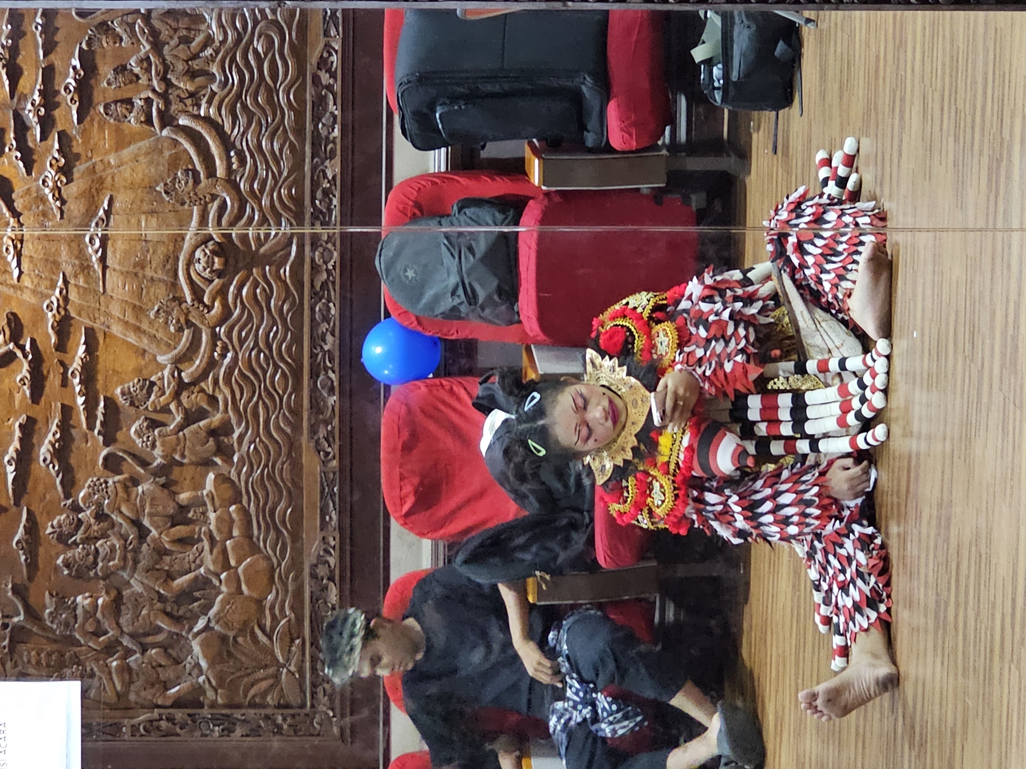 This media file captures a cultural performance or event in Denpasar, Indonesia. The scene appears to be indoors, possibly a theater or performance hall, with wooden floors and red seating. A central focus is a young person in elaborate traditional costume, characterized by a red, white, and black patterned outfit, a decorative headdress, and brightly colored makeup. The performer is lying down or reclining, with eyes closed, perhaps in a moment of rest or during a specific part of the performance.
To the left of the performer, a blue balloon is visible, adding a touch of whimsy. In the background, there are seats that appear to be occupied by other individuals, some covered in black fabric. One person, wearing a dark outfit and a cap, is seated and looking downwards. Another is leaning back in a red chair.
To the far left, a large, intricately carved wooden panel is prominently featured. This carving depicts a scene with multiple figures and possibly mythological elements, suggesting a rich cultural heritage. The carving has a textured, detailed surface that contrasts with the smoother elements of the setting.
The lighting suggests an indoor environment, and no specific time of day or weather conditions are discernible. The overall atmosphere seems to be one of cultural expression and performance, with the performer's attire and makeup indicating a traditional dance or ritual. The presence of onlookers and the stage-like setting point to a public event. A small portion of white paper with some text is visible at the bottom left corner, but the text is illegible.