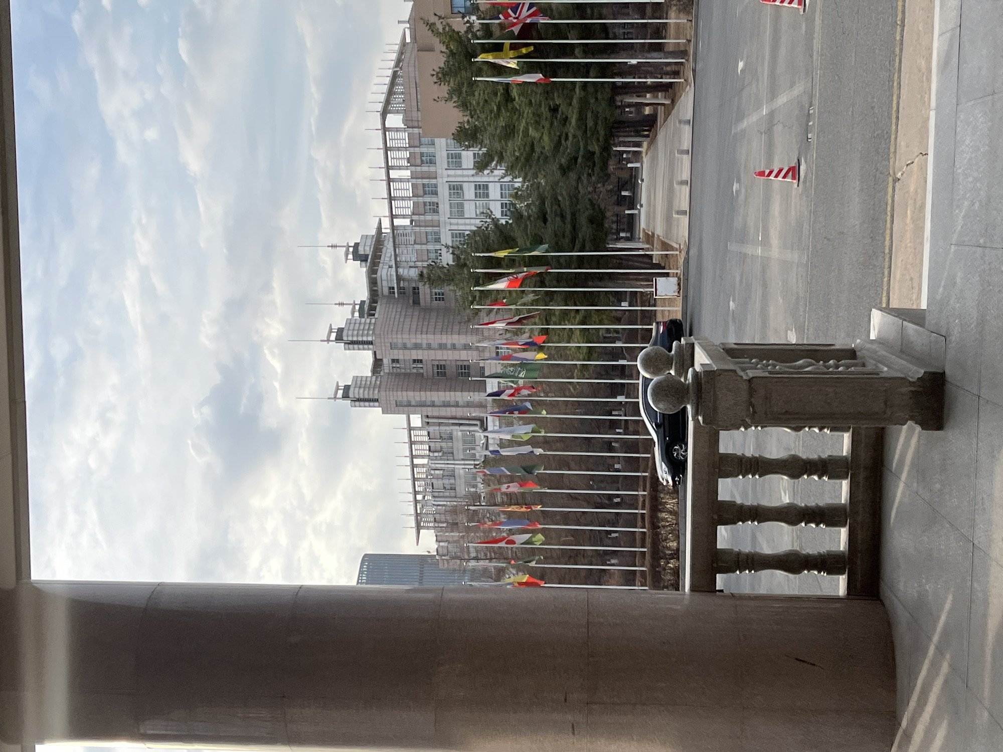The image shows a view of a street lined with flags from what appears to be an elevated position, possibly a building or structure with a concrete column and ornate railing in the foreground. The flags are on poles and arranged in rows along the sides of the street. Beyond the flags, there are buildings of varying heights and architectural styles. The sky is overcast with a mix of light and dark clouds. The location appears to be Changchun City, China, given the architectural style of the buildings and general environment. A dark-colored car is parked nearby on the street. Red and white traffic cones are present. The scene appears to be taken during the day.