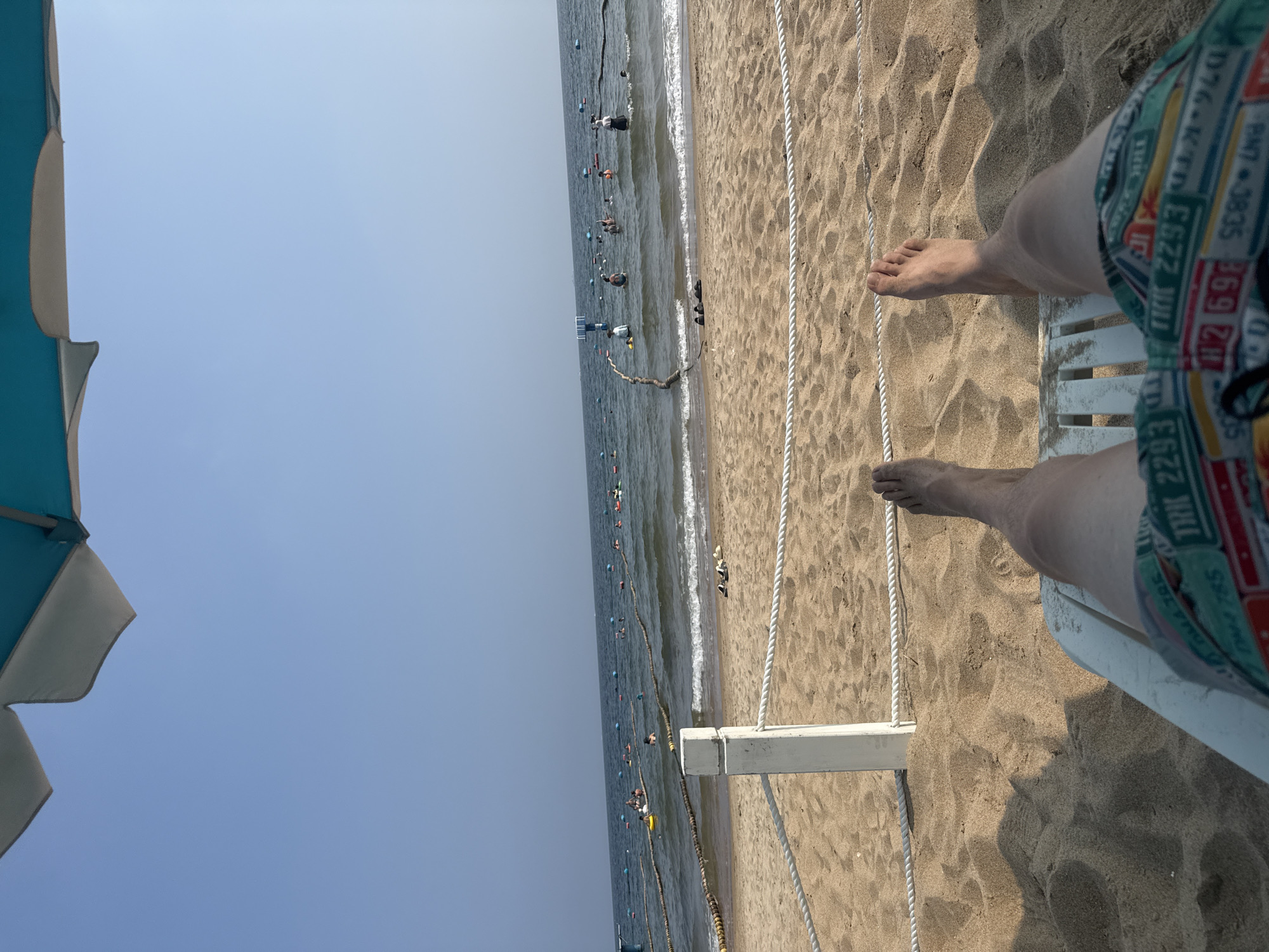 The image shows a first-person perspective of a person relaxing on a beach chair at a beach in Qinhuangdao, China. The foreground features the person's legs and feet, clad in colorful swim trunks, resting on a white plastic beach chair. The sand is light brown and textured with small ripples. A white wooden post with a thick white rope cordon stretches across the sand in front of the chair.
Beyond the sandy area, the ocean is visible, with gentle waves rolling towards the shore. Several people are in the water, some swimming and others standing. Buoys and ropes mark designated swimming areas in the sea. The sky is clear and a pale blue, with the edge of a beach umbrella visible in the upper part of the frame. The overall impression is a sunny day at the beach.