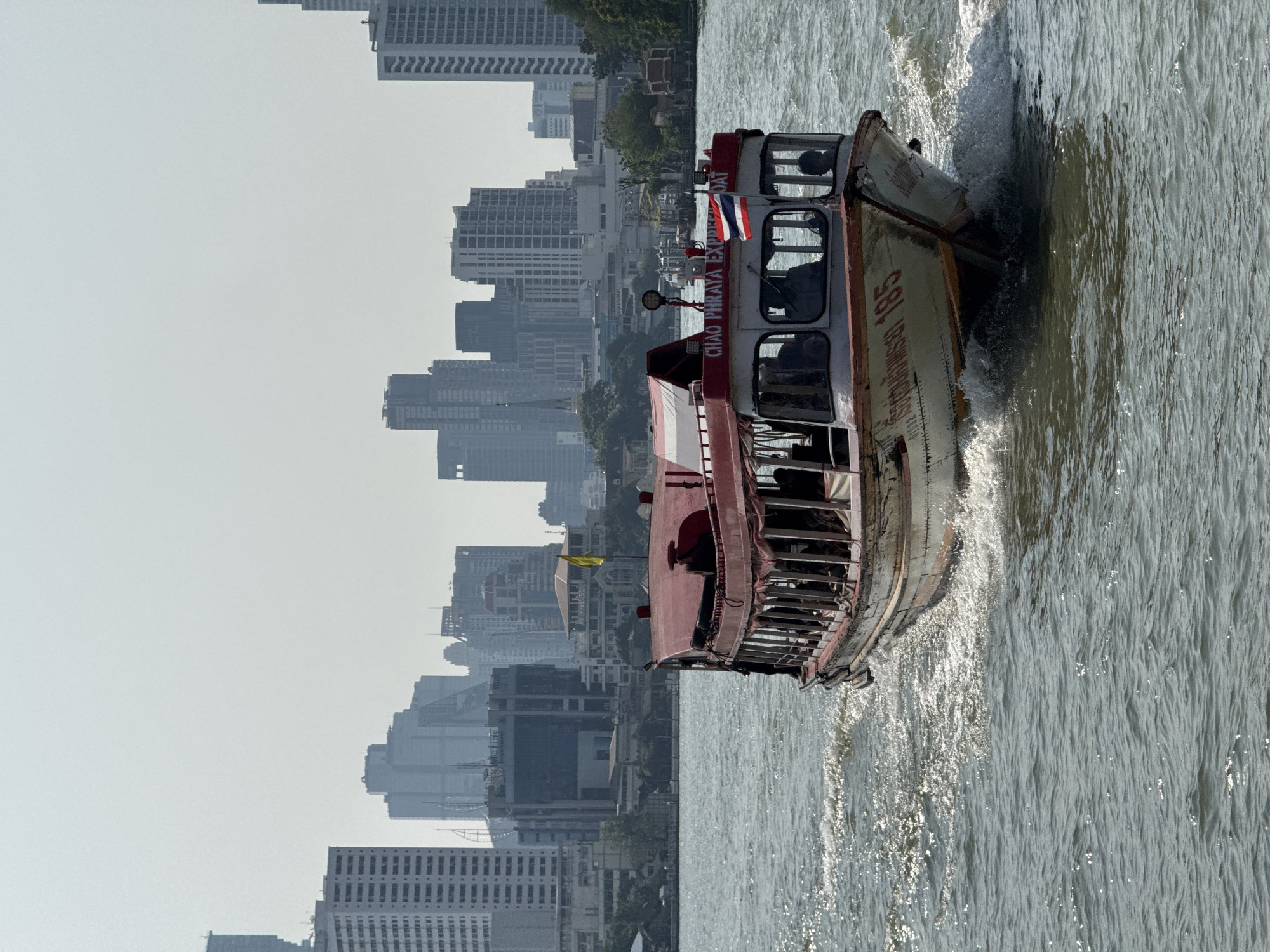 A weathered white and red "Chao Phraya Express Boat 185" is depicted in motion on the Chao Phraya River in Bangkok, Thailand. The vessel, identifiable by "CHAO PHRAYA EXPRESS BOAT" and the number "185" written in Thai script and numerals on its bow, is traveling from right to left across the frame, creating a discernible foamy wake behind its stern and spray at its bow. A Thai national flag is affixed to the front section of the boat's red upper roof structure. Multiple dark, silhouetted figures are visible inside the open-sided passenger compartment, and at least two individuals are present in the enclosed front cockpit area, one appearing to be at the helm. The river water is dark, choppy, and shows glints of sunlight reflecting on its surface. The background features a sprawling urban skyline of Bangkok, comprising numerous high-rise buildings and skyscrapers under a pale, hazy sky, partially obscuring the distant structures. Green foliage and other structures, including one with a yellow flag, are visible along the riverbanks.