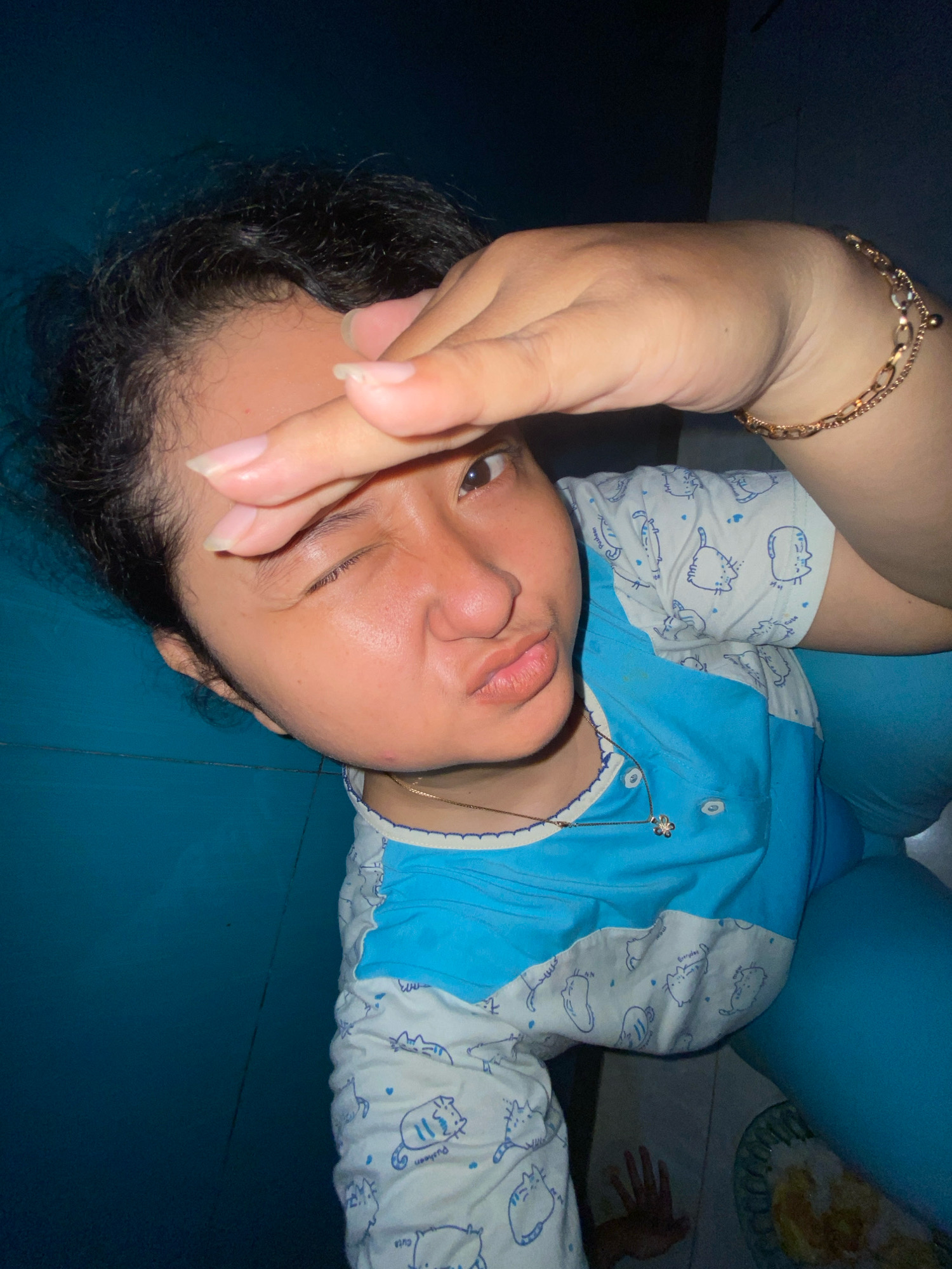 The image shows a close-up, high-angle shot of a young person indoors, likely in Gelam, Indonesia. The person is lying down, covering their eyes with their right hand, and making a pouting expression. They are wearing a blue and white patterned pajama set with cartoon cat designs. A thin gold chain with a flower pendant is visible around their neck, and a gold bracelet adorns their right wrist. The background consists of a dark blue surface, possibly a wall or furniture, and a hint of a tiled floor with a plate and a hand visible in the lower right corner. The lighting is dim, suggesting an indoor setting at night or in low light conditions.