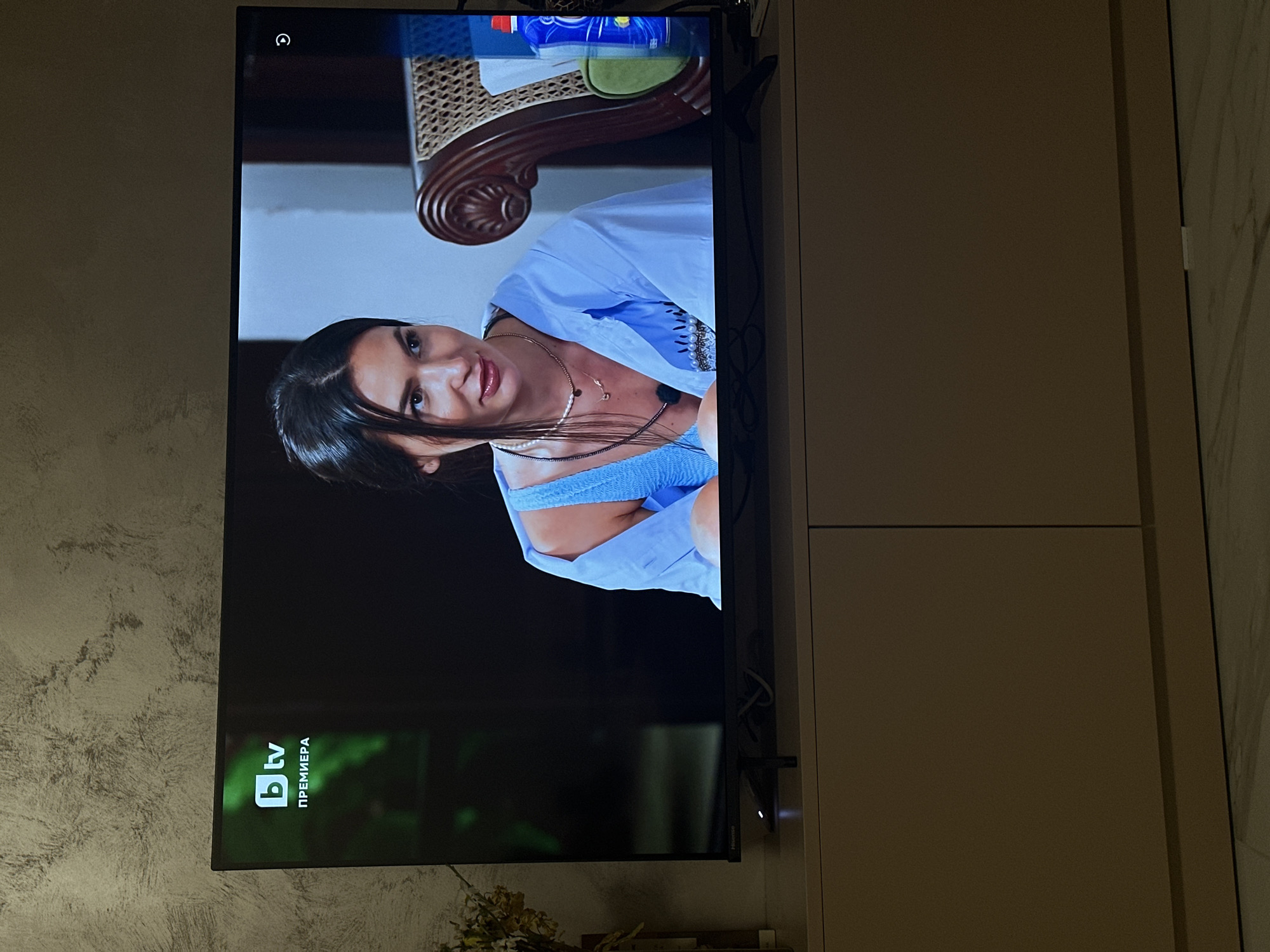The image shows a television screen displaying a woman in a blue shirt. The TV is mounted on a wall with light-colored cabinets. In the bottom left corner of the screen, there is a logo with the letters "bTV" and the word "ПРЕМИЕРА" (Premiere). Based on the logo, it is likely that the location is in Bulgaria. BTV is a television station that is based in Sofia, Bulgaria.