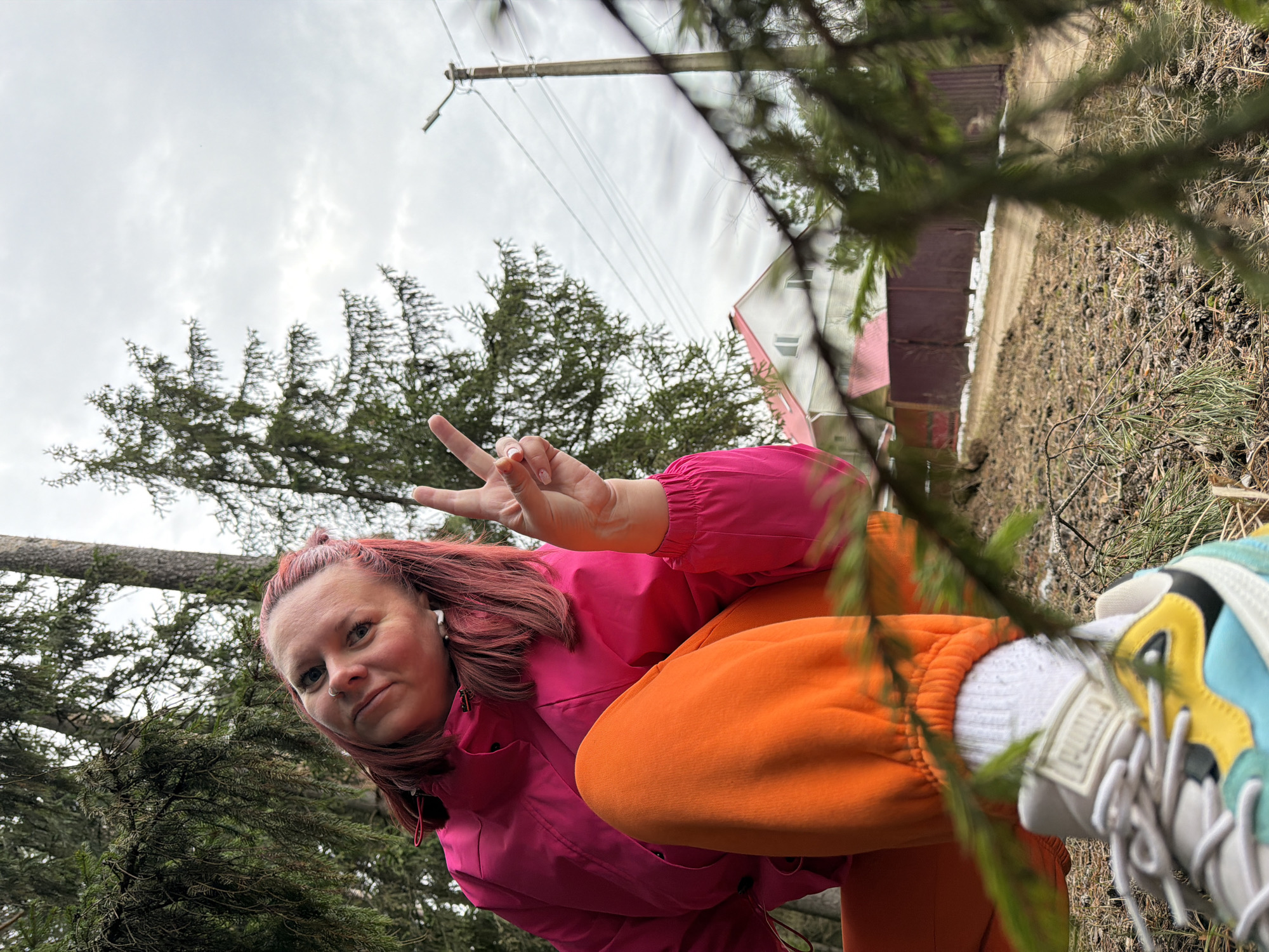 A woman with reddish-brown hair, wearing a pink jacket and orange sweatpants, is giving the peace sign outdoors. She has a white earbud in her left ear. The woman is surrounded by trees and under an overcast sky. In the background, there is a building with a red roof and white siding. A power pole and lines are visible in the upper portion of the image. A shoe with a Puma logo is also visible. The image appears to be taken at an angle. Given the location prompt, the scene is in Chaikovsky, Russia.