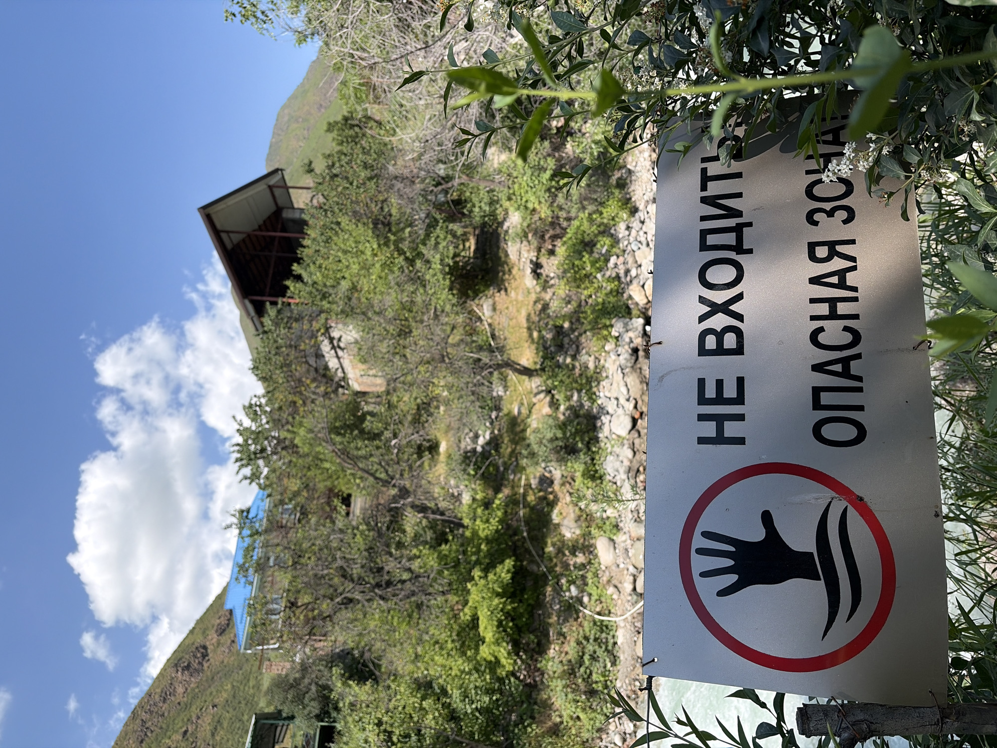 In an outdoor scene with lush green vegetation and a bright blue sky dotted with white clouds, a metal sign is prominently displayed in the foreground. The sign features a red circle with a black silhouette of a hand emerging from water, indicating danger. Below this symbol, text in black Cyrillic letters reads "НЕ ВХОДИТЬ" (Do not enter) on the top line and "ОПАСНАЯ ЗОНА" (Danger zone) on the bottom line. The background reveals a mountainous landscape with buildings nestled among trees and hills. The overall environment suggests a natural setting near a body of water, possibly a river or stream, given the imagery on the sign.
