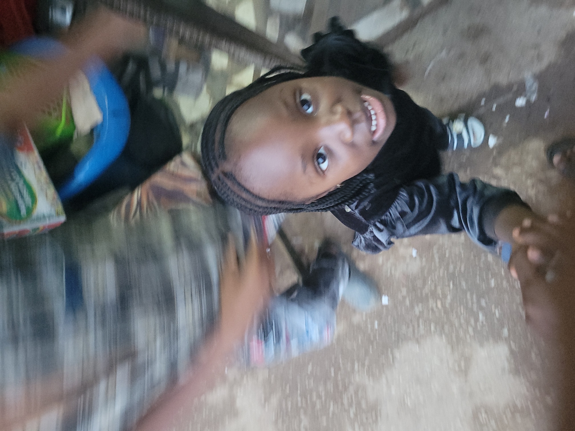 A young, smiling girl with braided hair looks up into the camera. She wears a dark-colored top. The photo is taken from a high angle, making her appear smaller. The background is blurry. A hand holds her hand in the bottom-right corner of the frame. The ground appears to be concrete or a similar surface with some debris scattered around. Other blurred objects are visible, but not distinctly discernible. The location appears to be an interior space, with possible urban elements suggesting Jos, Nigeria.