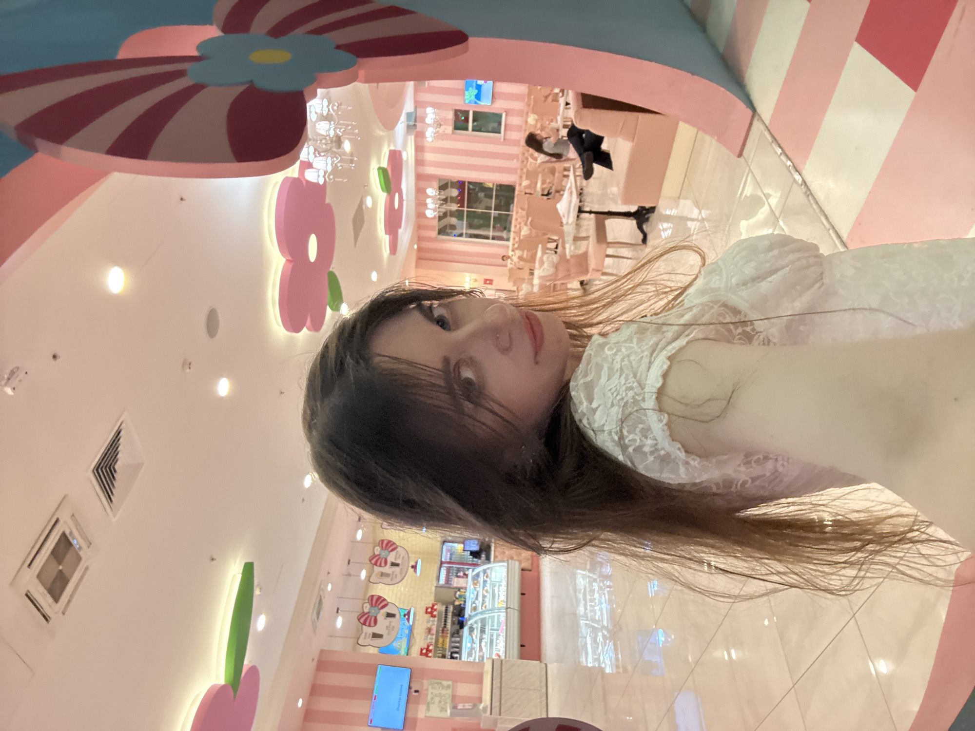 The image shows a woman in a white lace top taking a selfie in a brightly lit, heavily themed interior. The decor features pastel pink and white stripes, oversized flower and bow decorations on the walls and ceiling, and Hello Kitty motifs, suggesting it's a Hello Kitty-themed cafe. In the background, other customers are visible seated at tables. A display case filled with pastries and desserts is also visible. Based on the decor and theme, the location is likely the Hello Kitty Beauty Cafe in Moscow, Russia.